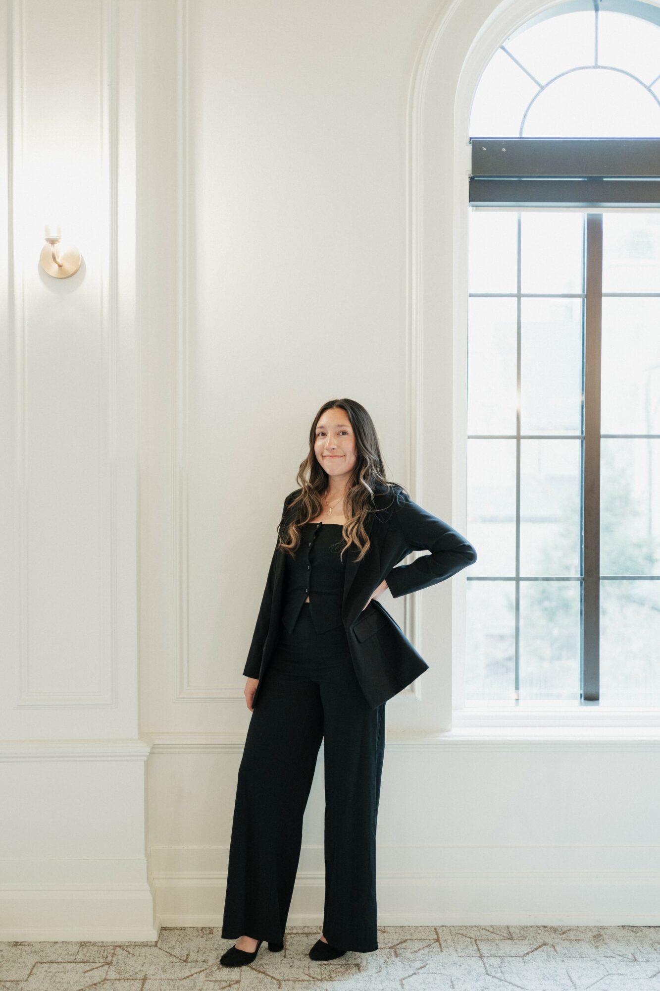 Woman in black suit standing indoors near large window with arched top, smiling, with hand on hip, light-colored walls, and carpeted floor.