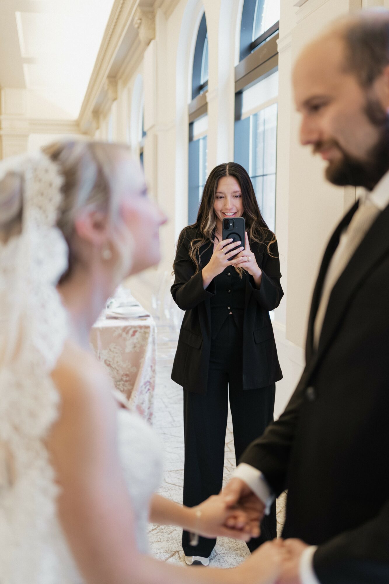 A bride and groom hold hands while a woman in the background takes a photo with her phone inside a bright, elegant room.