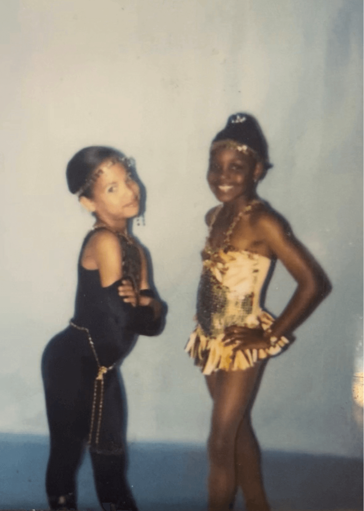 Two young girls in costumes stand side by side, smiling, against a plain background.