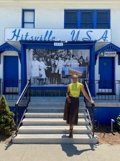 Woman in yellow top and dark skirt standing on stairs in front of a blue building with a sign reading 'Hitsville U.S.A.'