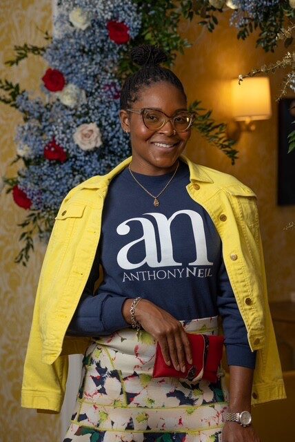 Woman with glasses smiling, wearing a yellow jacket over a navy shirt with white text, holding a red clutch, standing in front of floral arrangement.