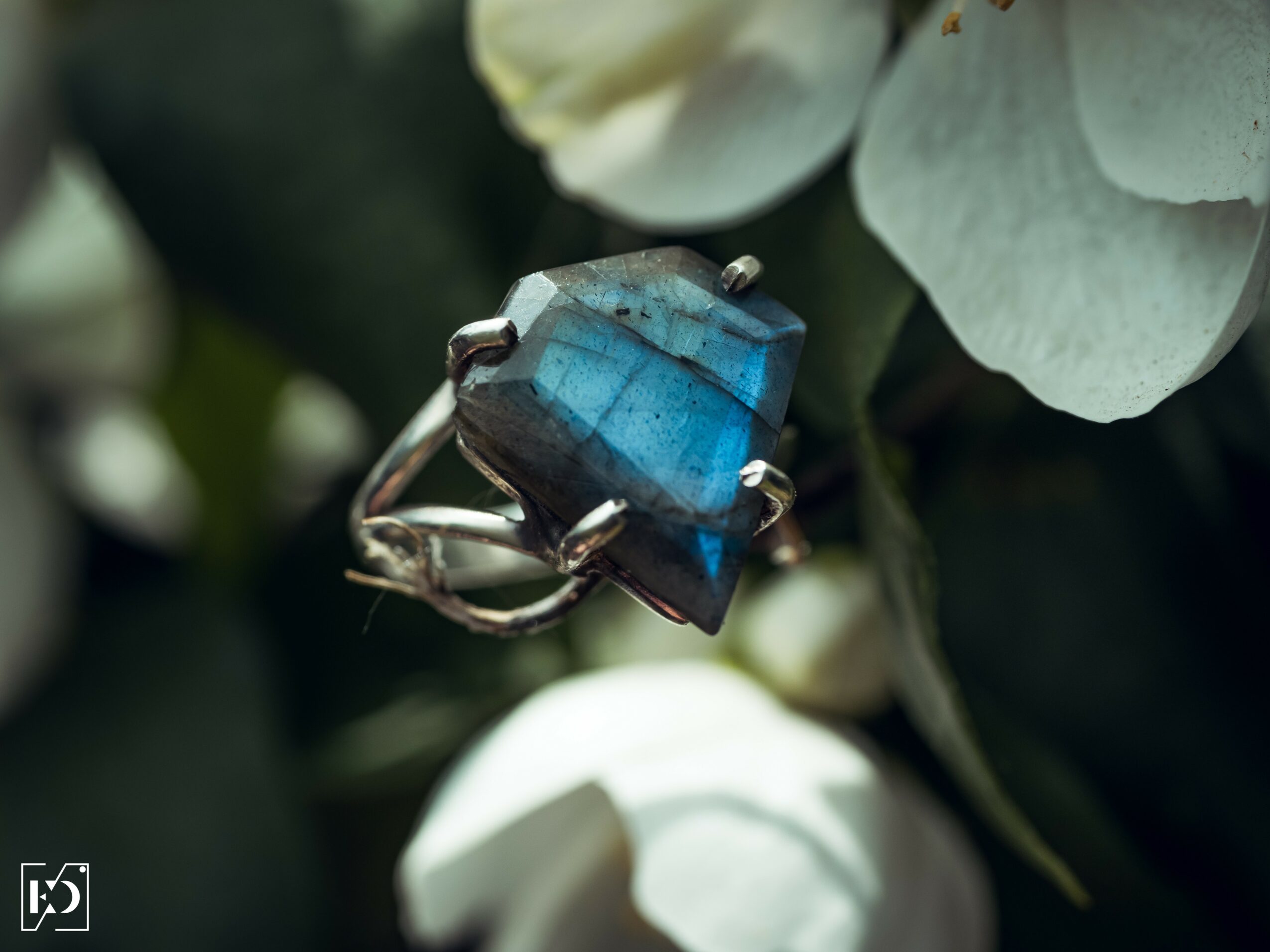 Ring with a large, square blue gemstone on a person's finger, surrounded by white flowers and green leaves.