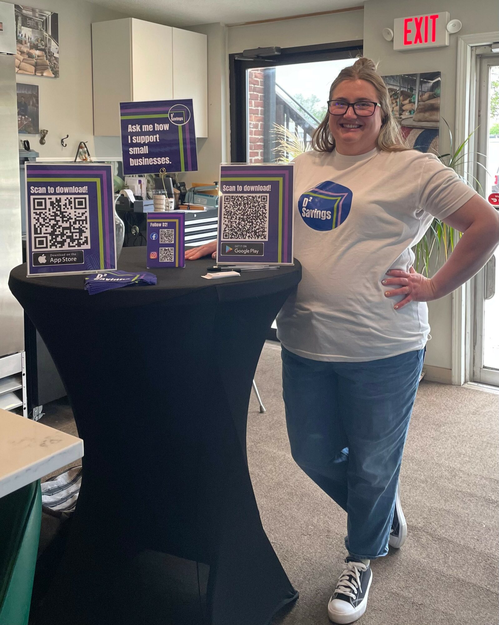 Woman standing next to a table with QR code signs inside a room with glass doors and windows, smiling with hands on hips.