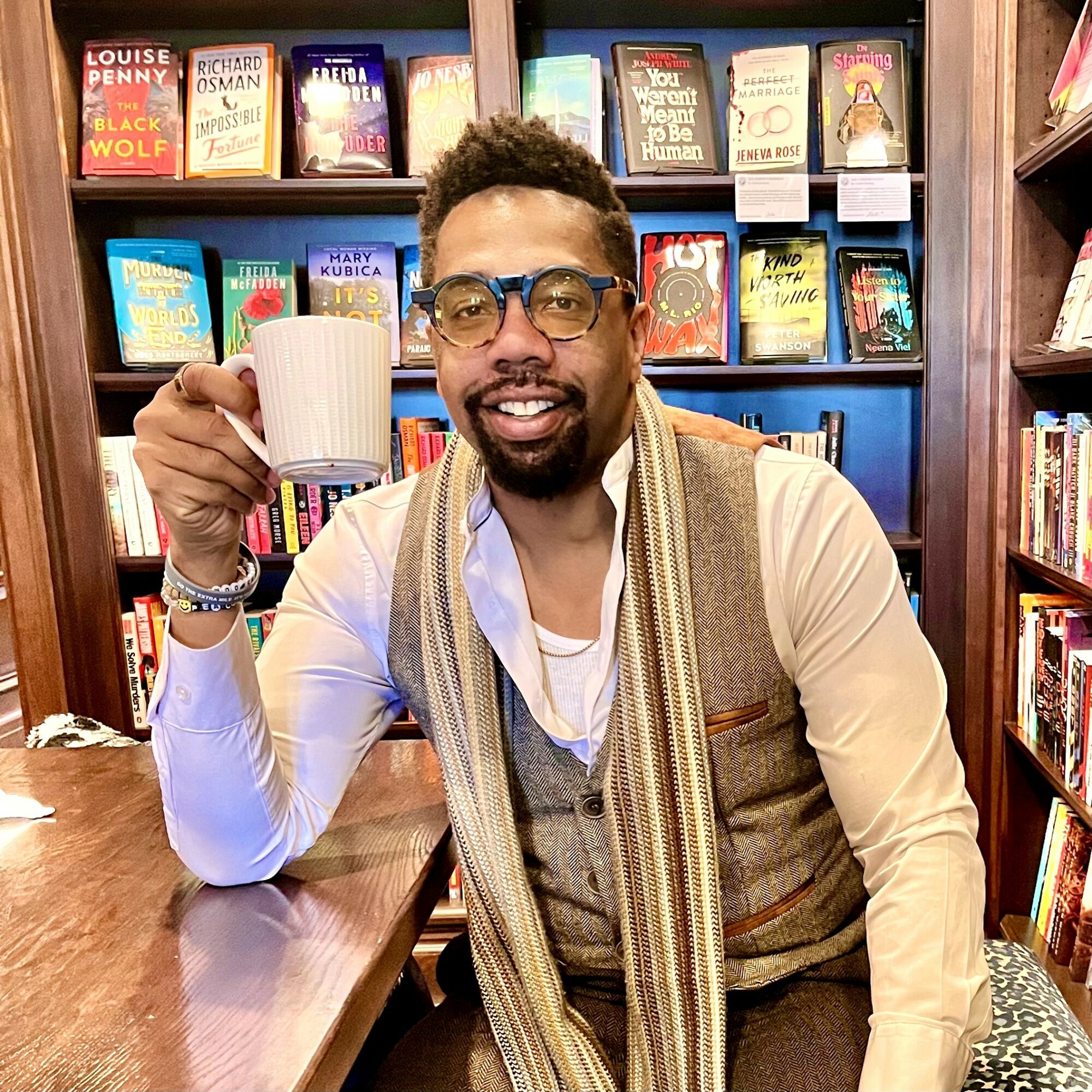Man with glasses and a beard smiling, holding a mug in a bookstore with shelves of books behind him.