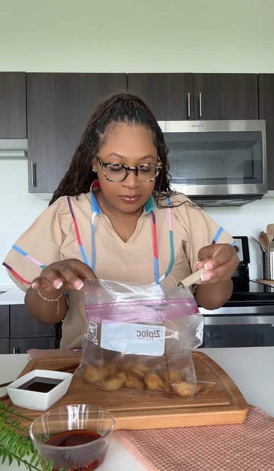 Woman with glasses and curly hair, wearing a beige shirt, is sealing a plastic bag with food inside in a kitchen.