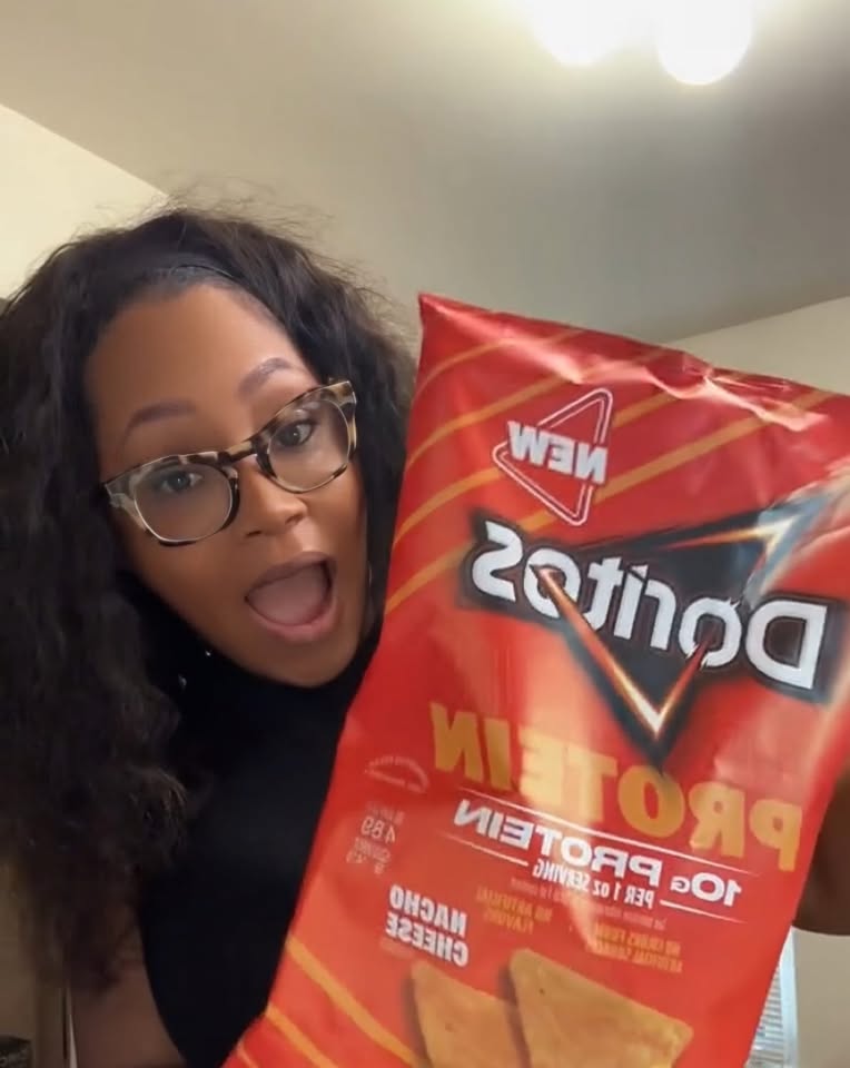 Person with glasses holding a bag of Doritos Nacho Cheese chips, smiling with mouth open, indoors.