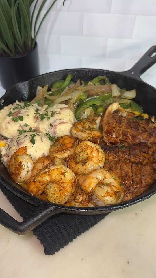 Cooked shrimp, chicken, and vegetables in a black skillet on a white surface, with a plant in the background.