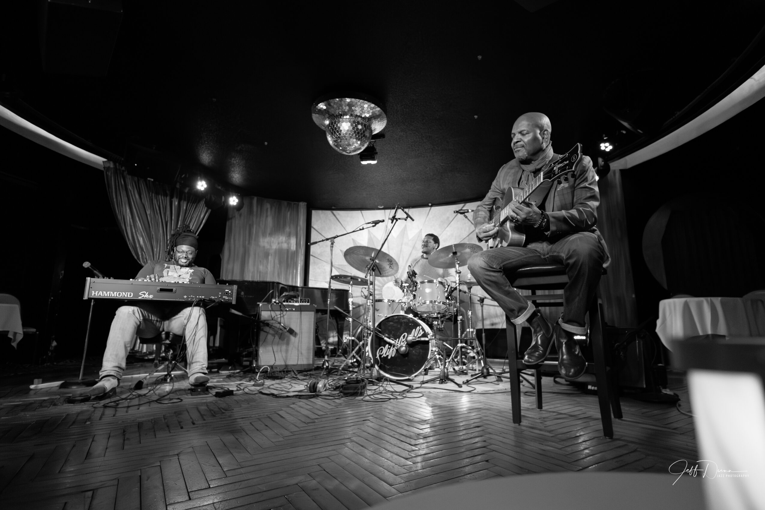 Three musicians perform on stage with instruments, including a keyboard, drums, and a seated singer, in a dimly lit venue.