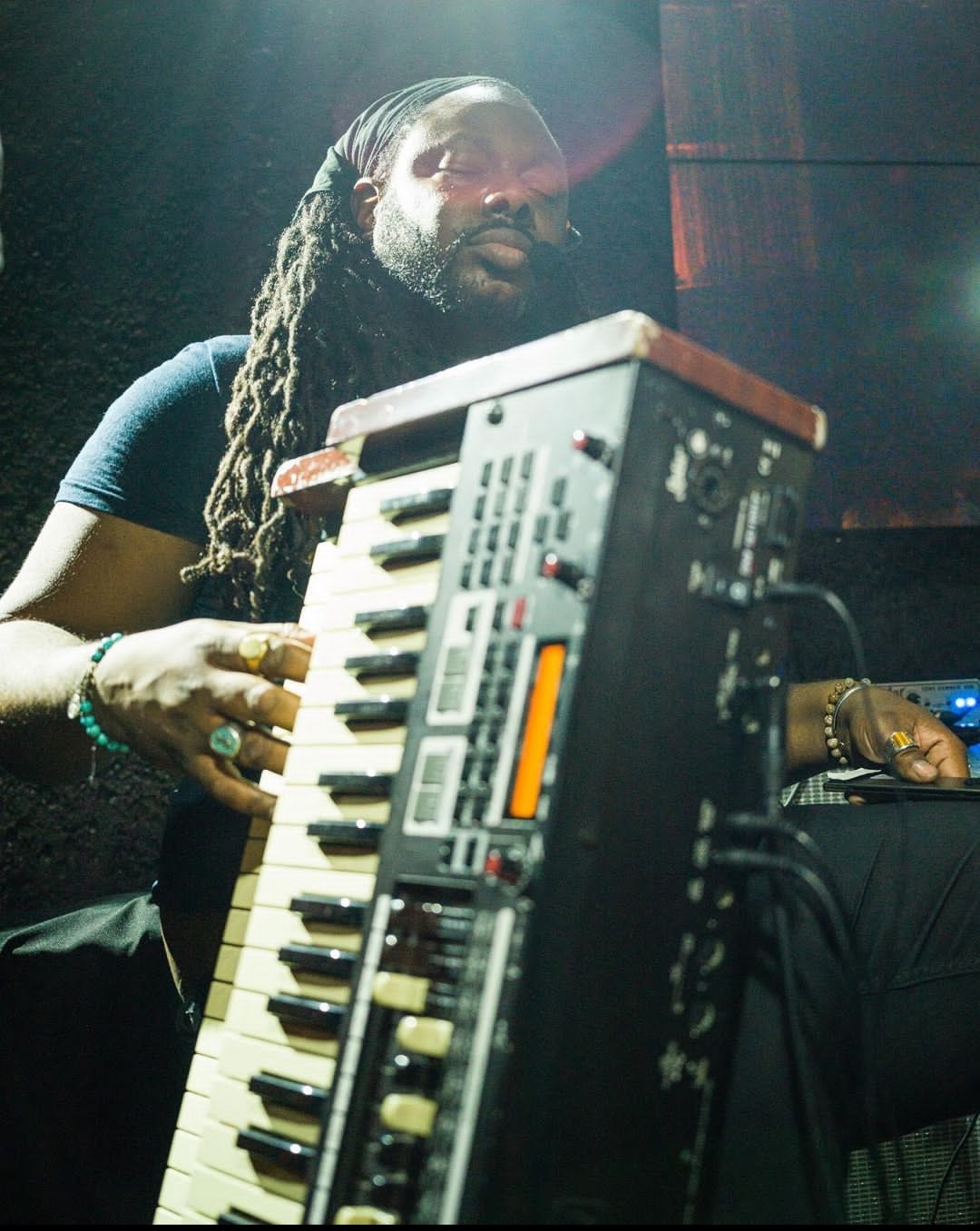 Person with long dreadlocks playing a keyboard, focused on music, in a dimly lit setting.