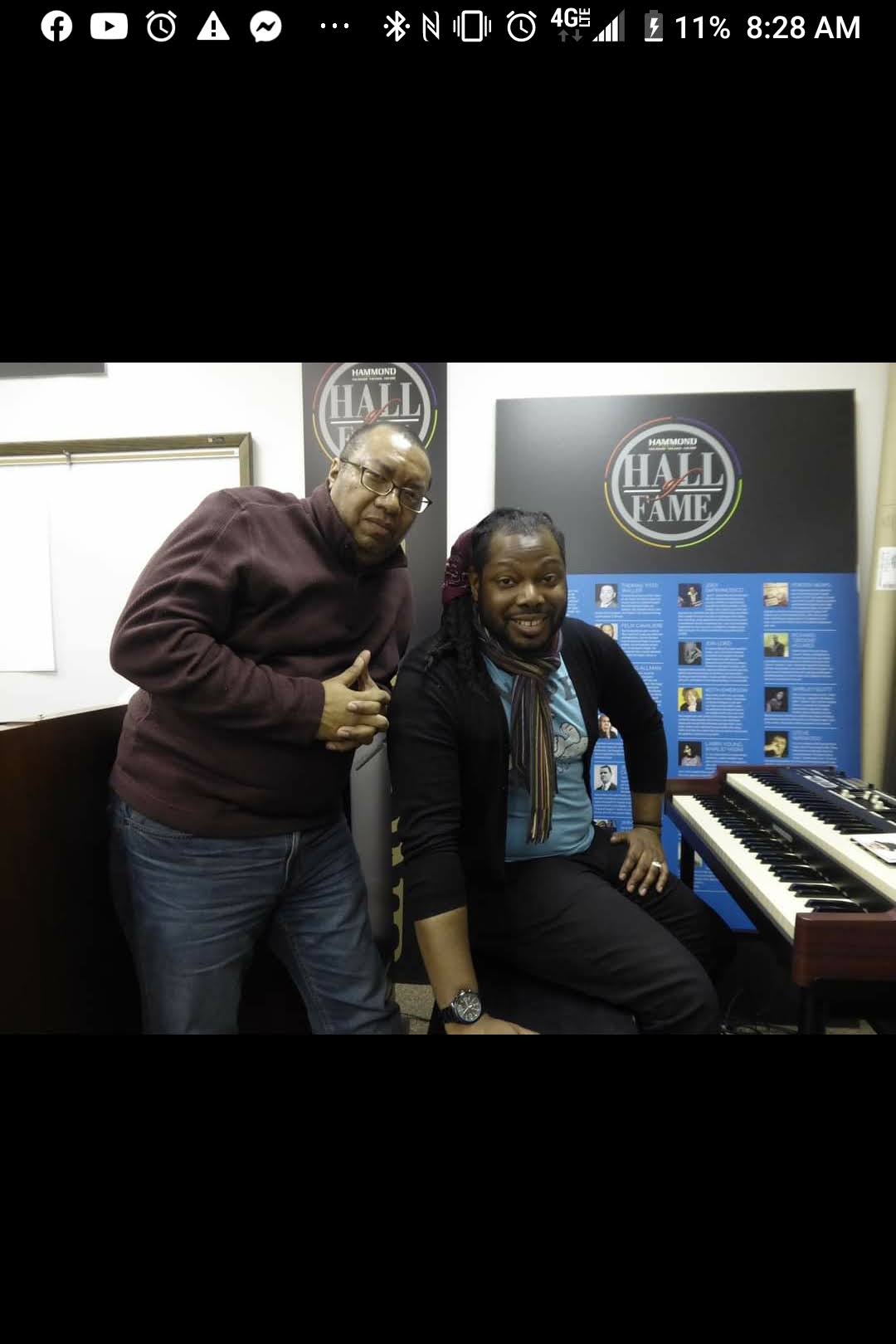 Two people in a music studio, one standing and one sitting at a keyboard, with music-related signs in the background.