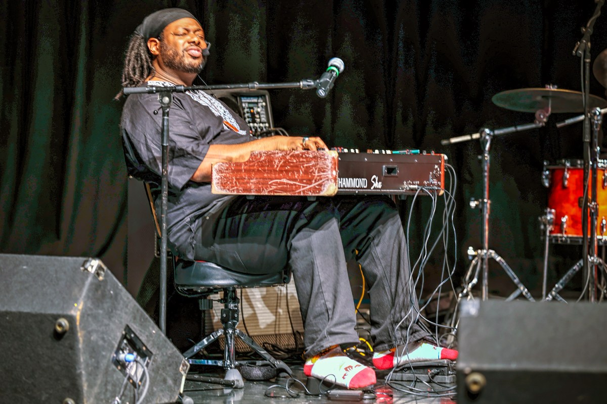 Musician playing keyboard on stage with drums in background, black curtain backdrop, seated, with microphone nearby.