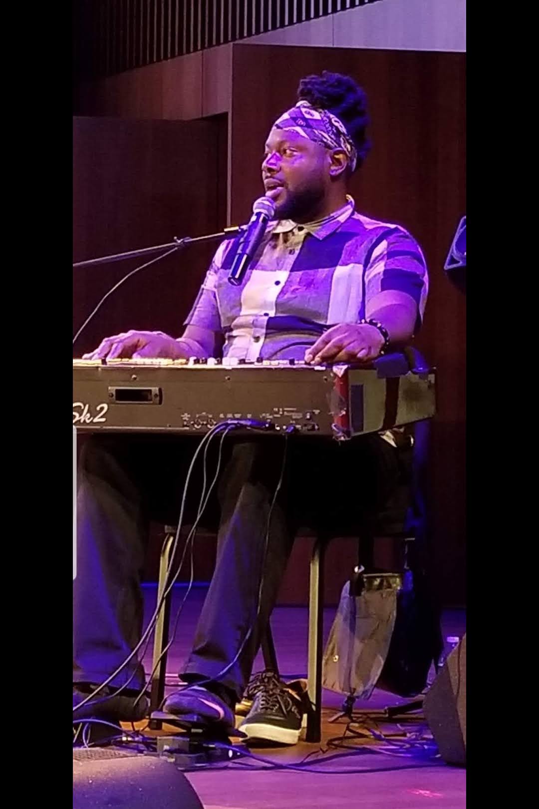 Person with glasses and headwrap playing keyboard on stage, singing into microphone, with dark background.