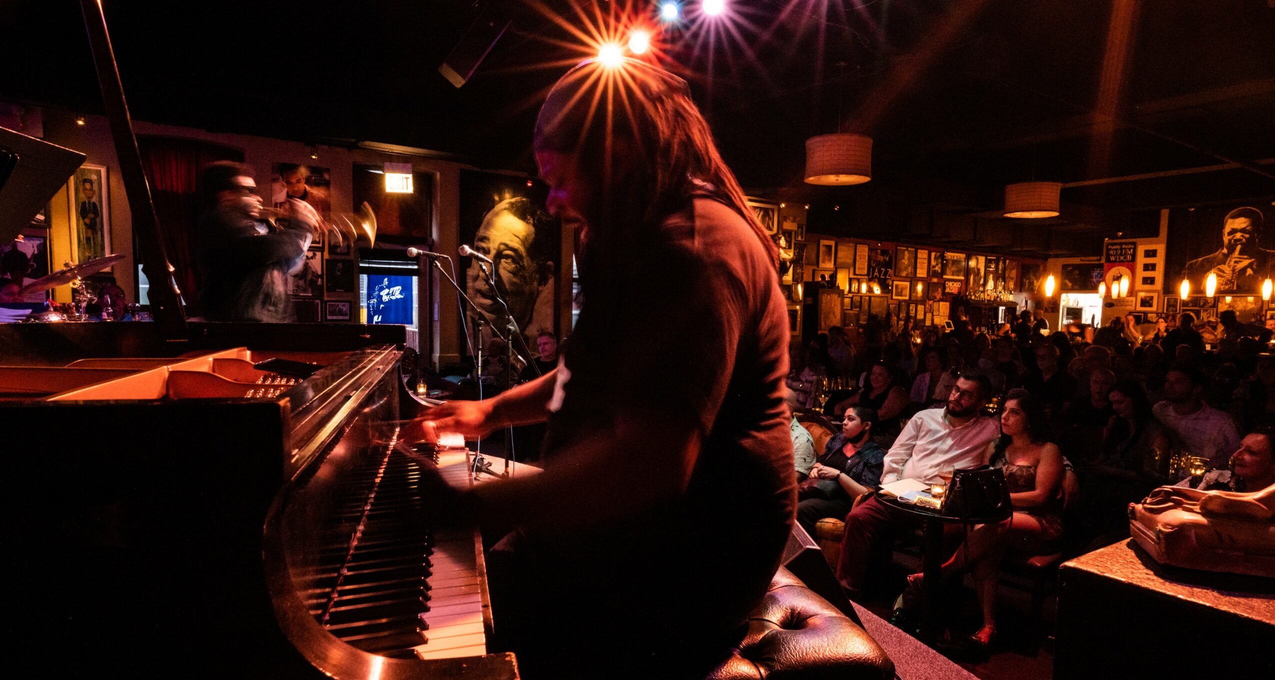 Person playing piano on stage with an audience in a dimly lit venue, colorful lights overhead, and people watching.