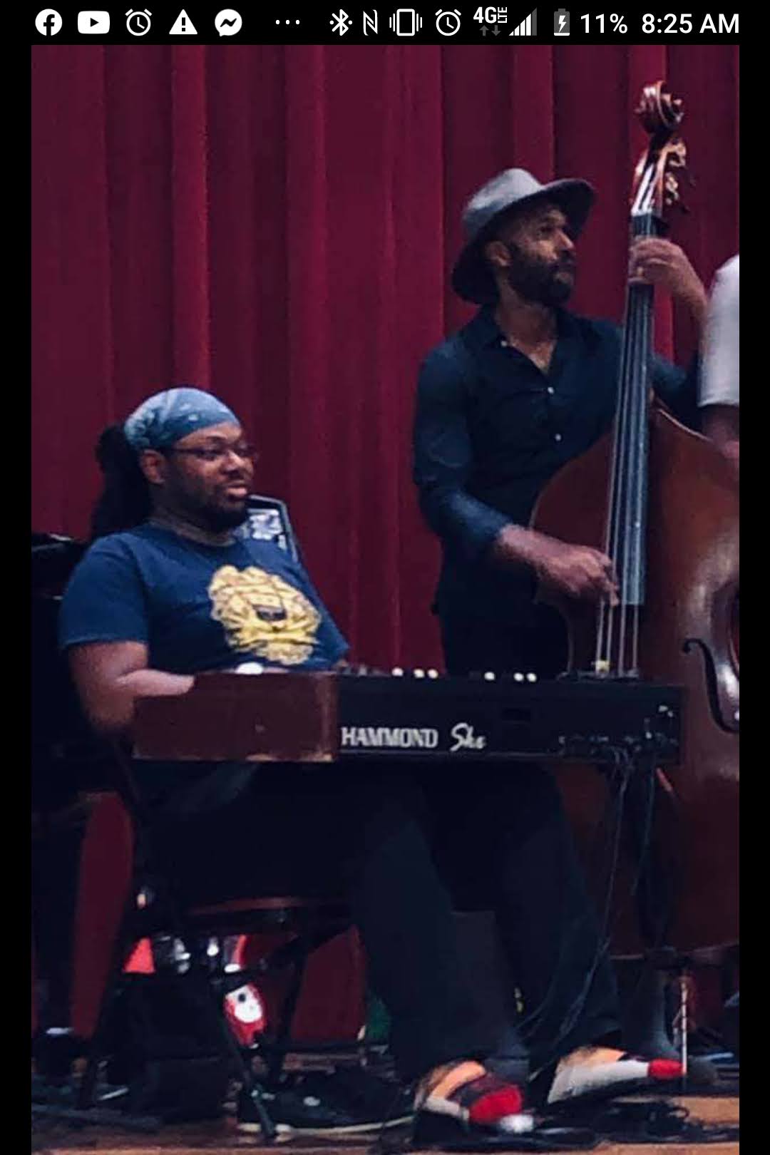 Two musicians perform on stage with a red curtain background; one plays keyboard, the other plays double bass.