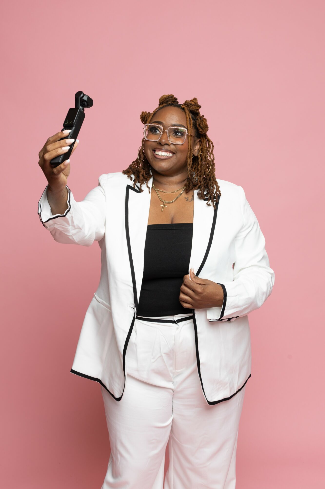 Woman with glasses and styled hair taking a selfie against pink background, smiling, wearing a white blazer and black top.