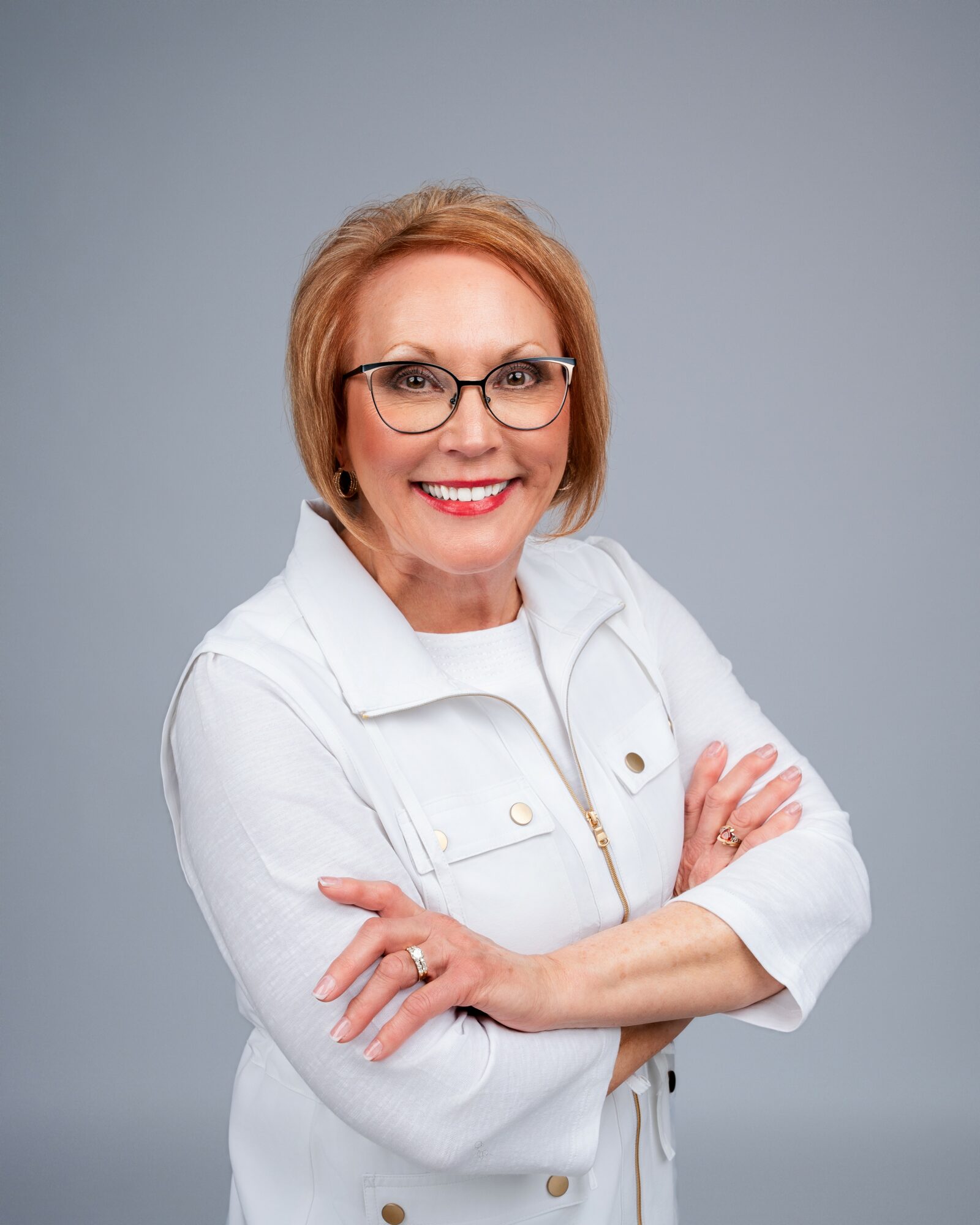 Woman with short red hair and glasses smiling, arms crossed, wearing a white jacket against a gray background.