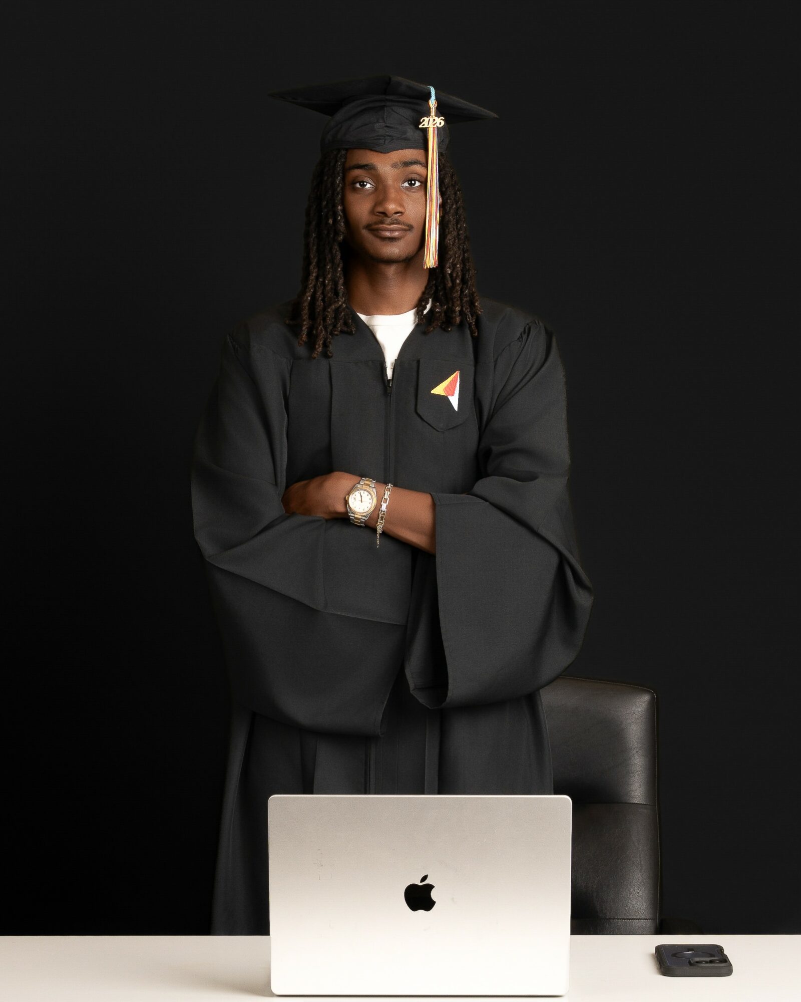 Graduate in cap and gown standing with arms crossed, in front of a laptop and a phone, against a black background.