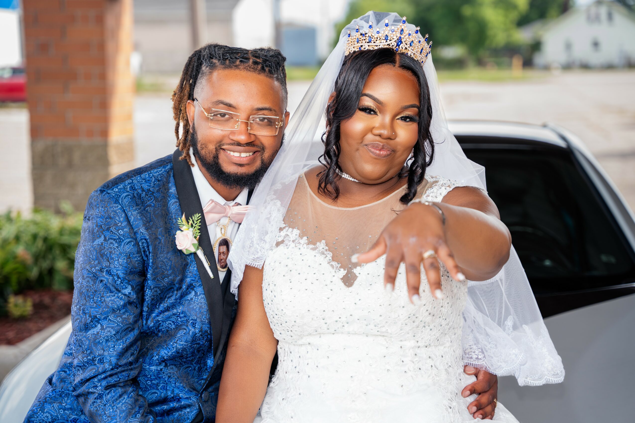 Bride and groom smiling outdoors, bride showing her ring, bride wearing a white wedding dress and veil, groom in a blue suit.