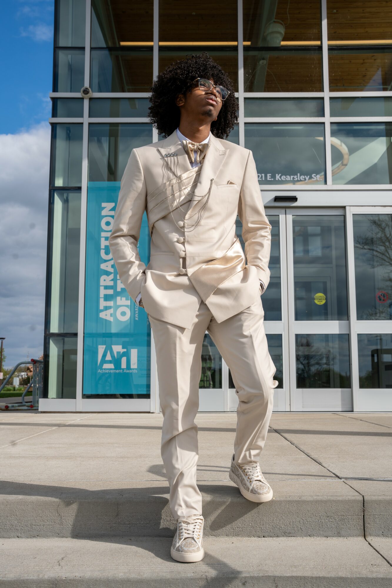 Young person with curly hair in a beige suit and sneakers standing outside a modern glass building.
