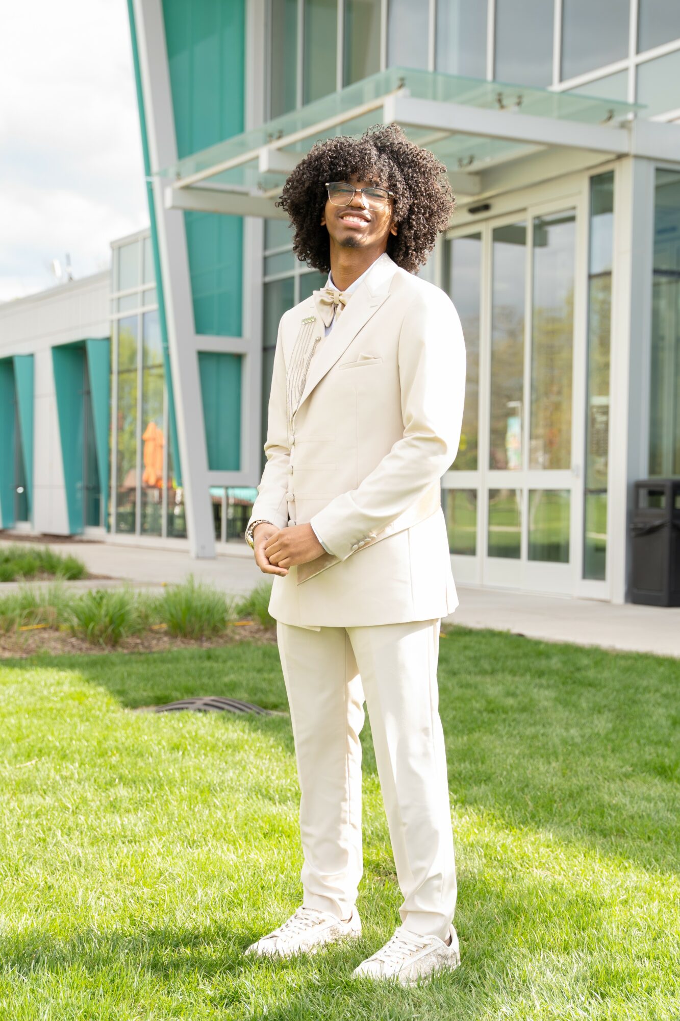Woman with curly hair in a white suit standing outdoors in front of a modern glass building, smiling.