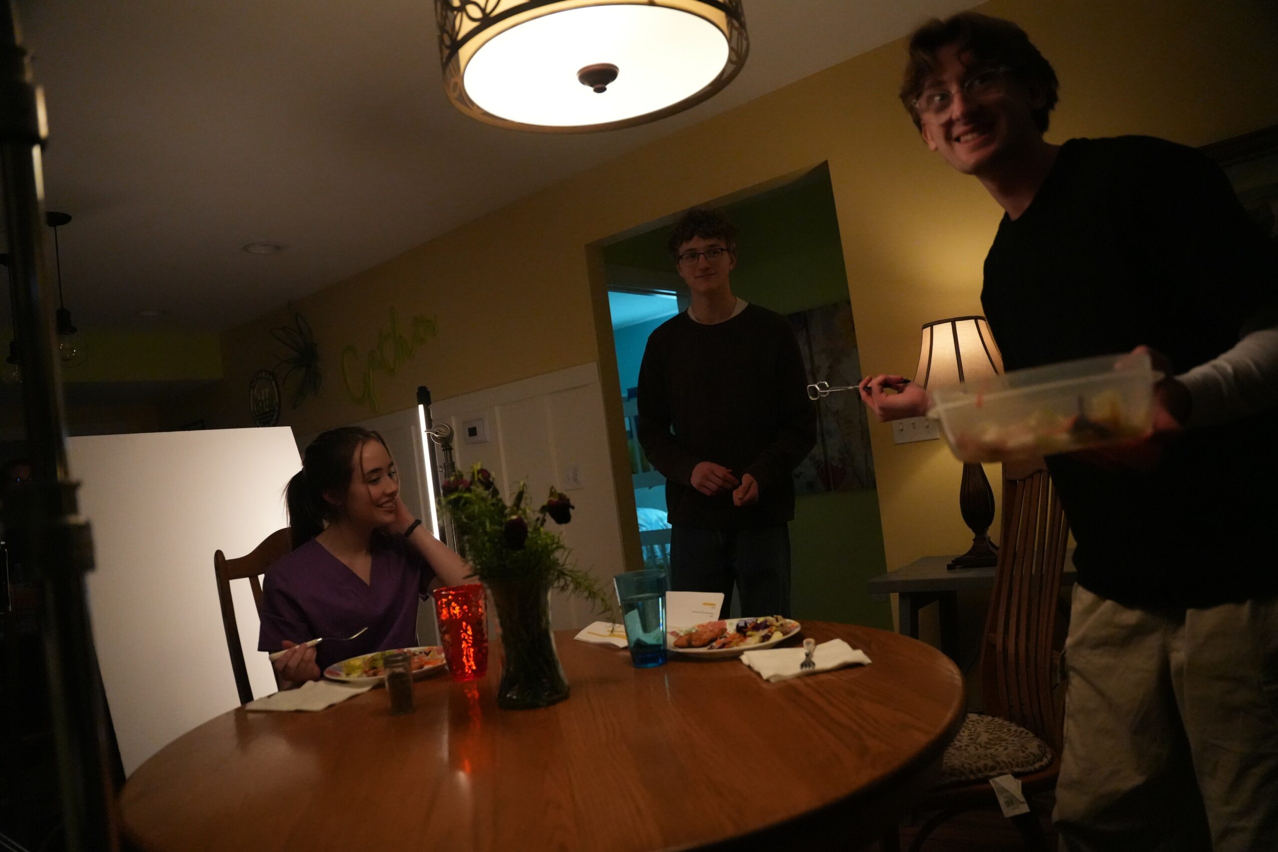 Three people in a dimly lit room, one sitting at a table, two standing, with drinks and food, smiling and talking.