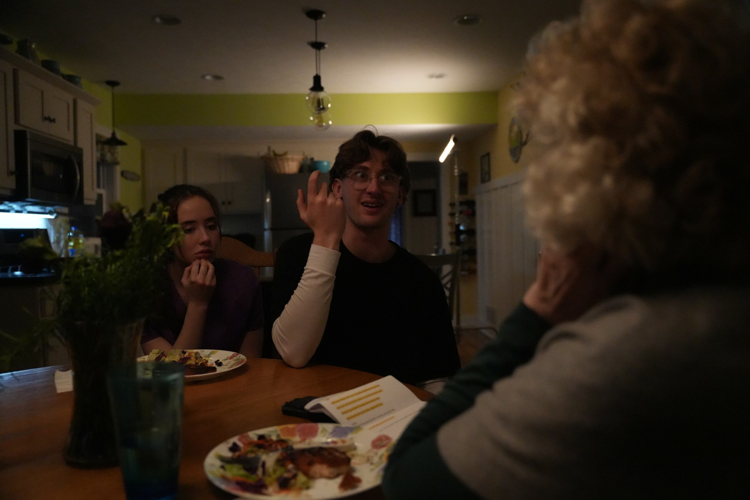 Three people sitting at a kitchen table engaged in conversation, with food and a notebook on the table.