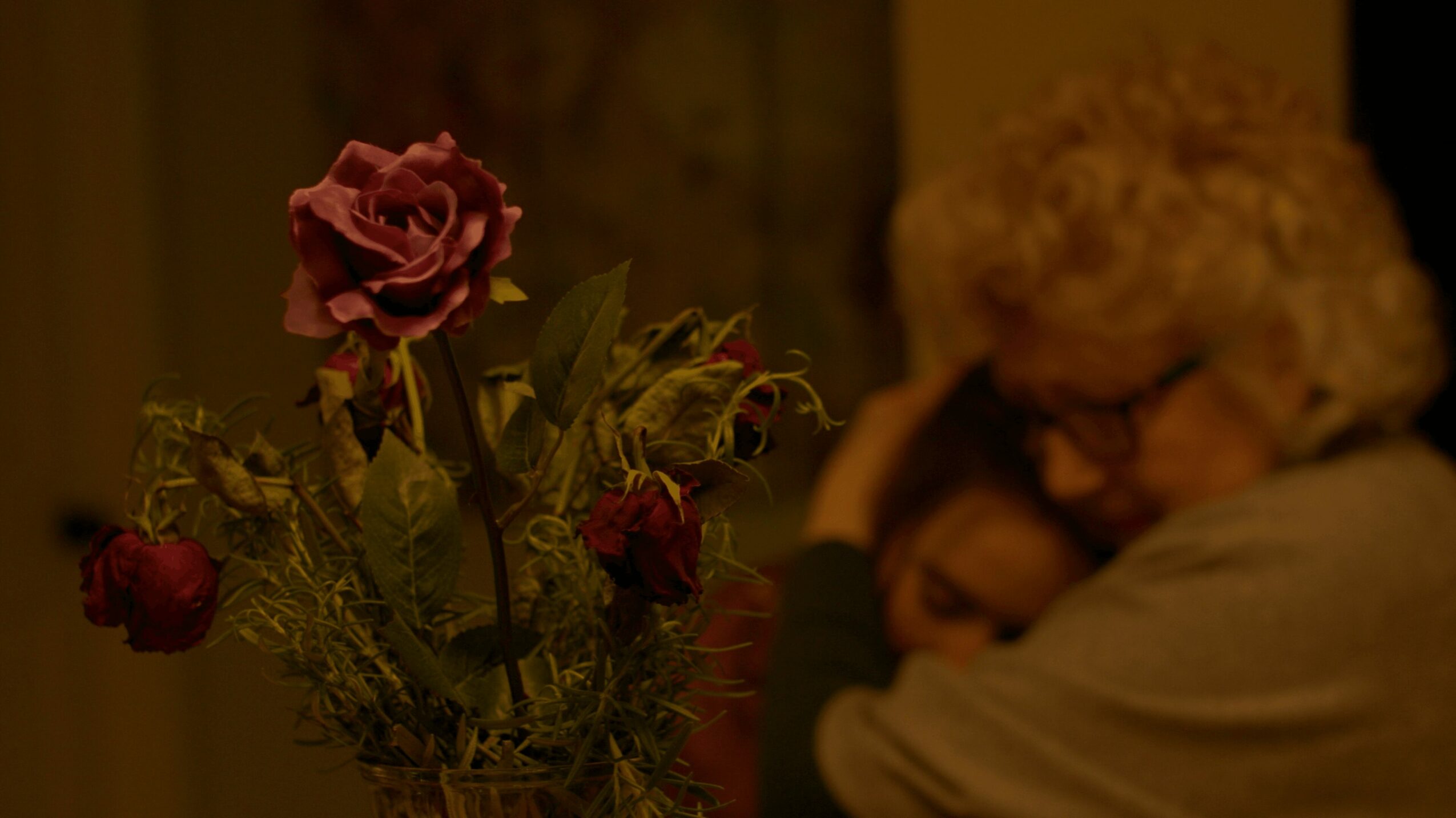 A person with curly hair and glasses hugs another person, with a bouquet of flowers nearby, in a warm setting.