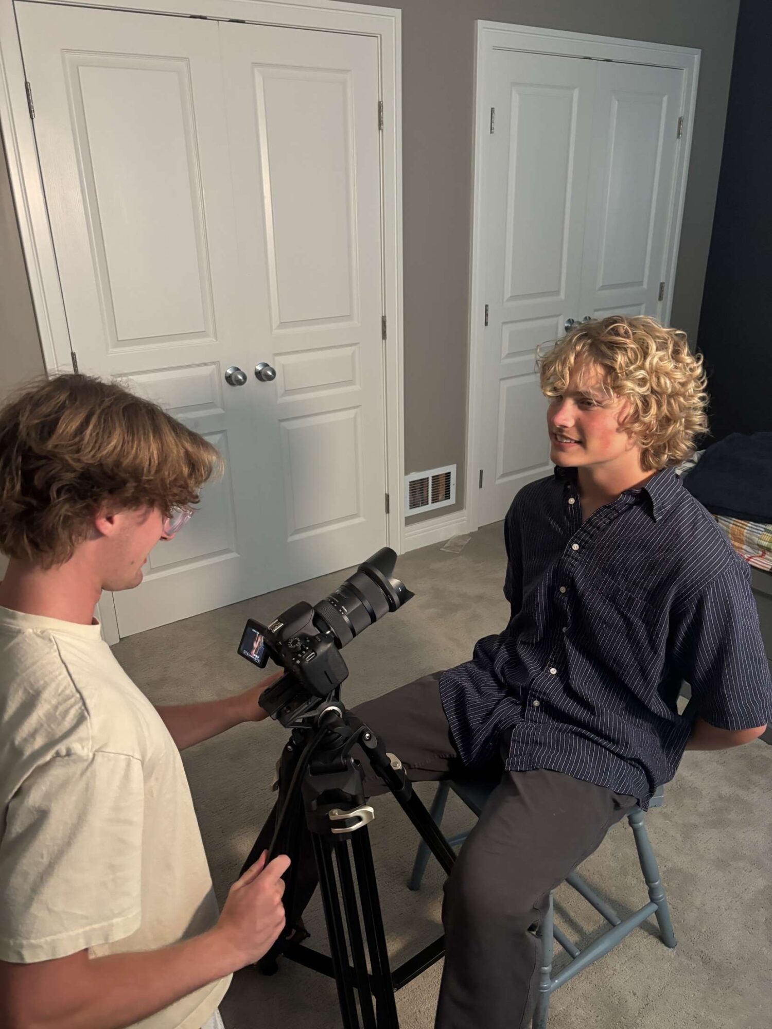 Two young men in a room with closed white doors, one sitting on a stool and the other holding a camera on a tripod, engaging in conversation.