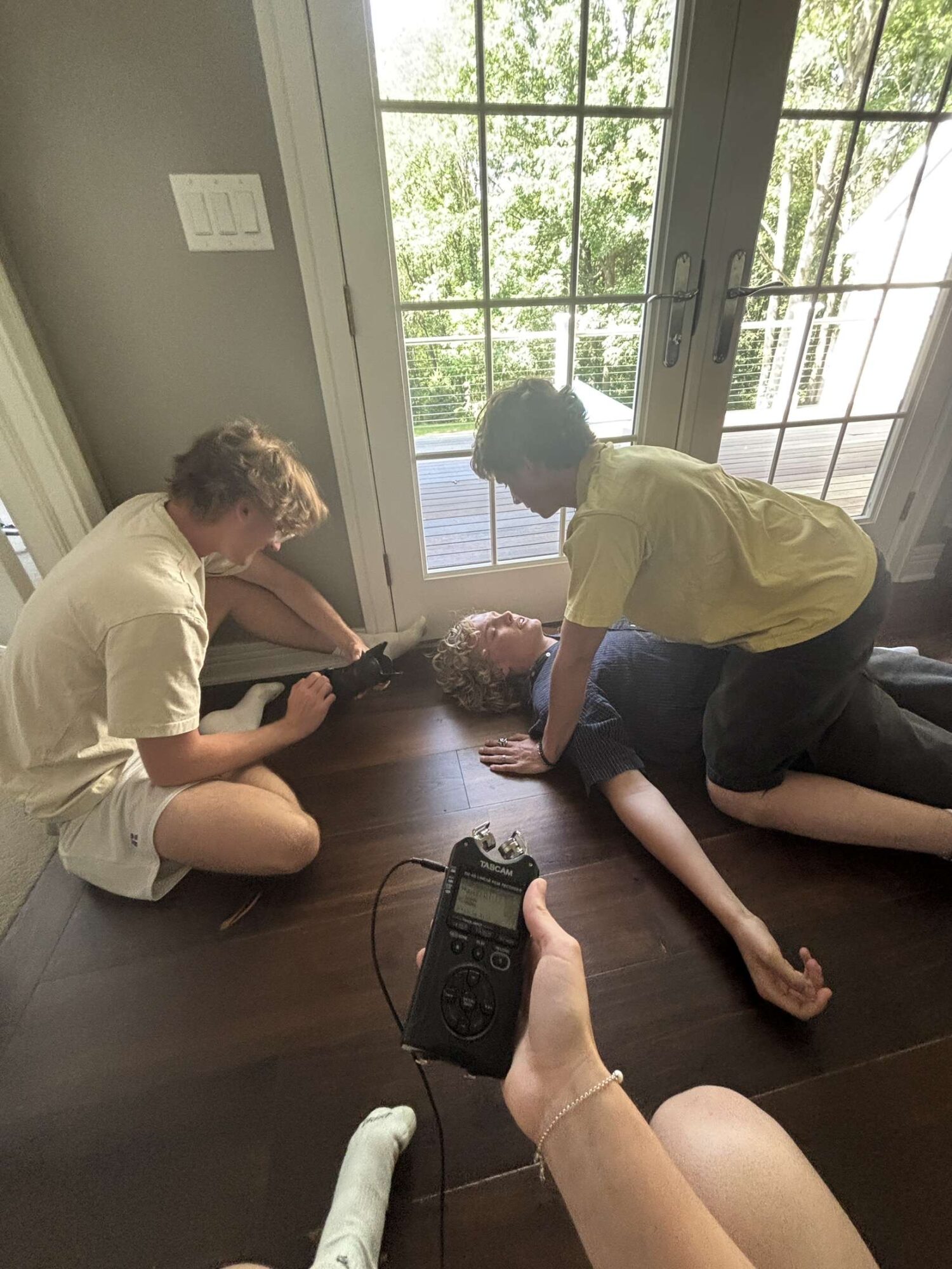 Two children are on the floor near a glass door, one reaching toward a bouquet of flowers, with a hand holding a remote control in the foreground.