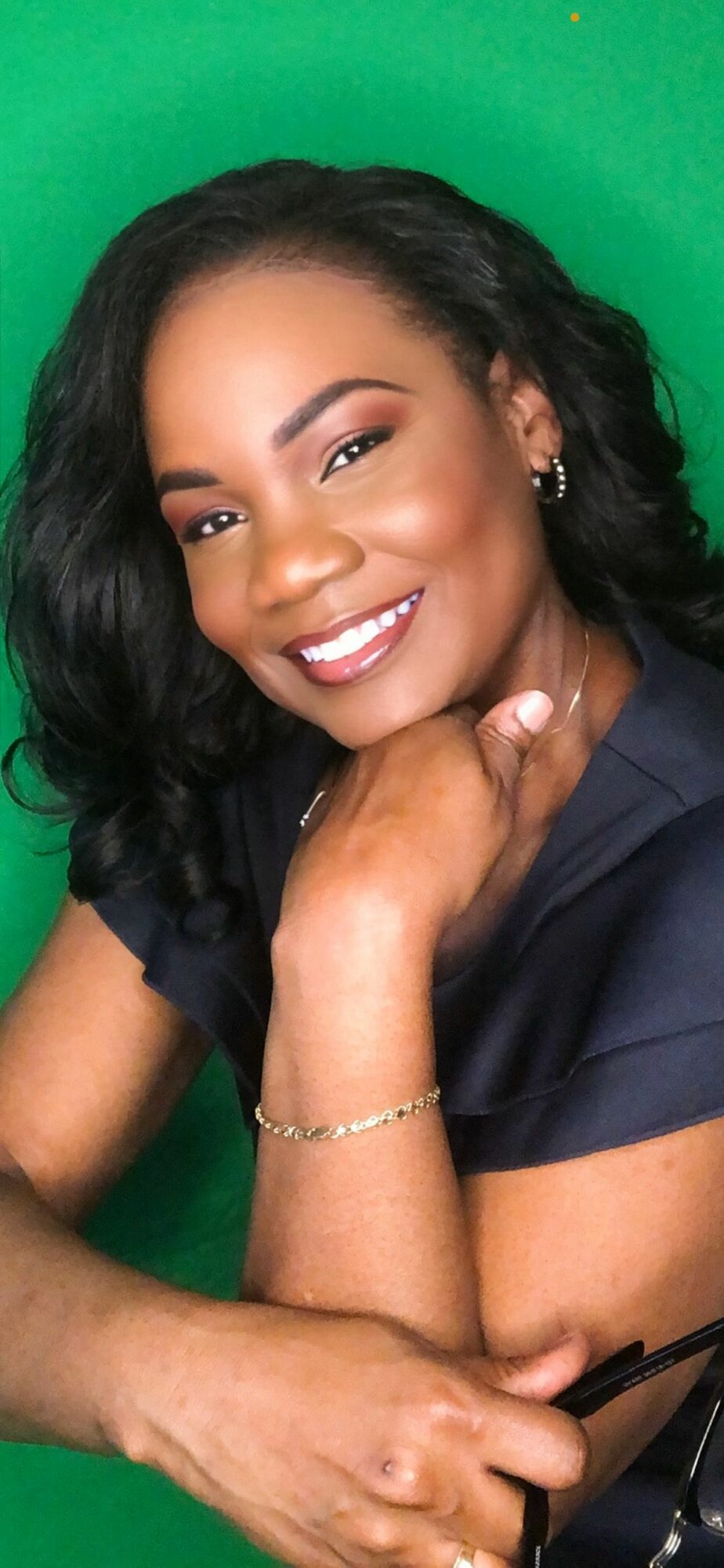 Smiling woman with dark curly hair and earrings, resting her chin on her hand, against a green background.