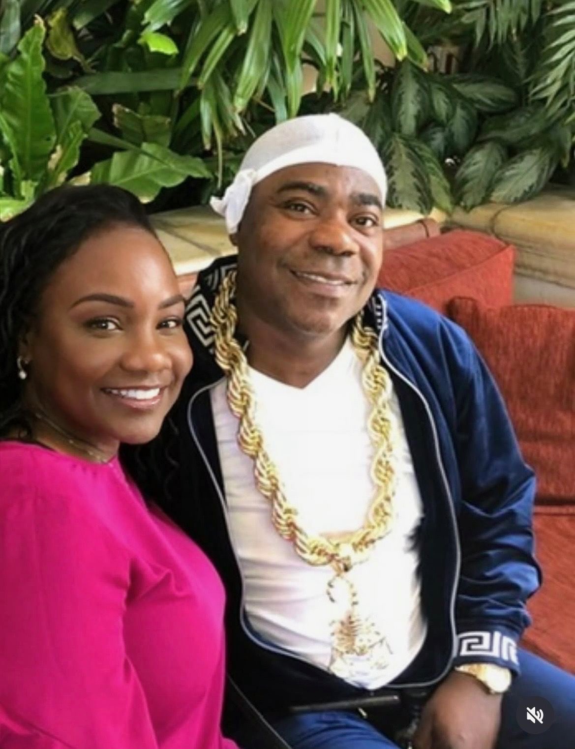 Two people smiling, one wearing a pink top and the other in a white head covering and gold jewelry, with green plants in background.