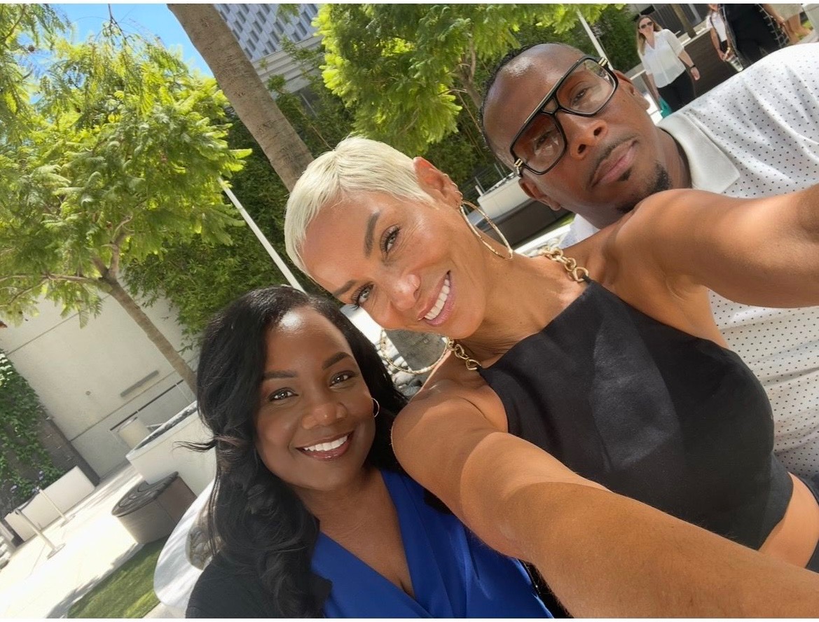 Three people smiling outdoors, two women and one man, with trees and a building in the background.