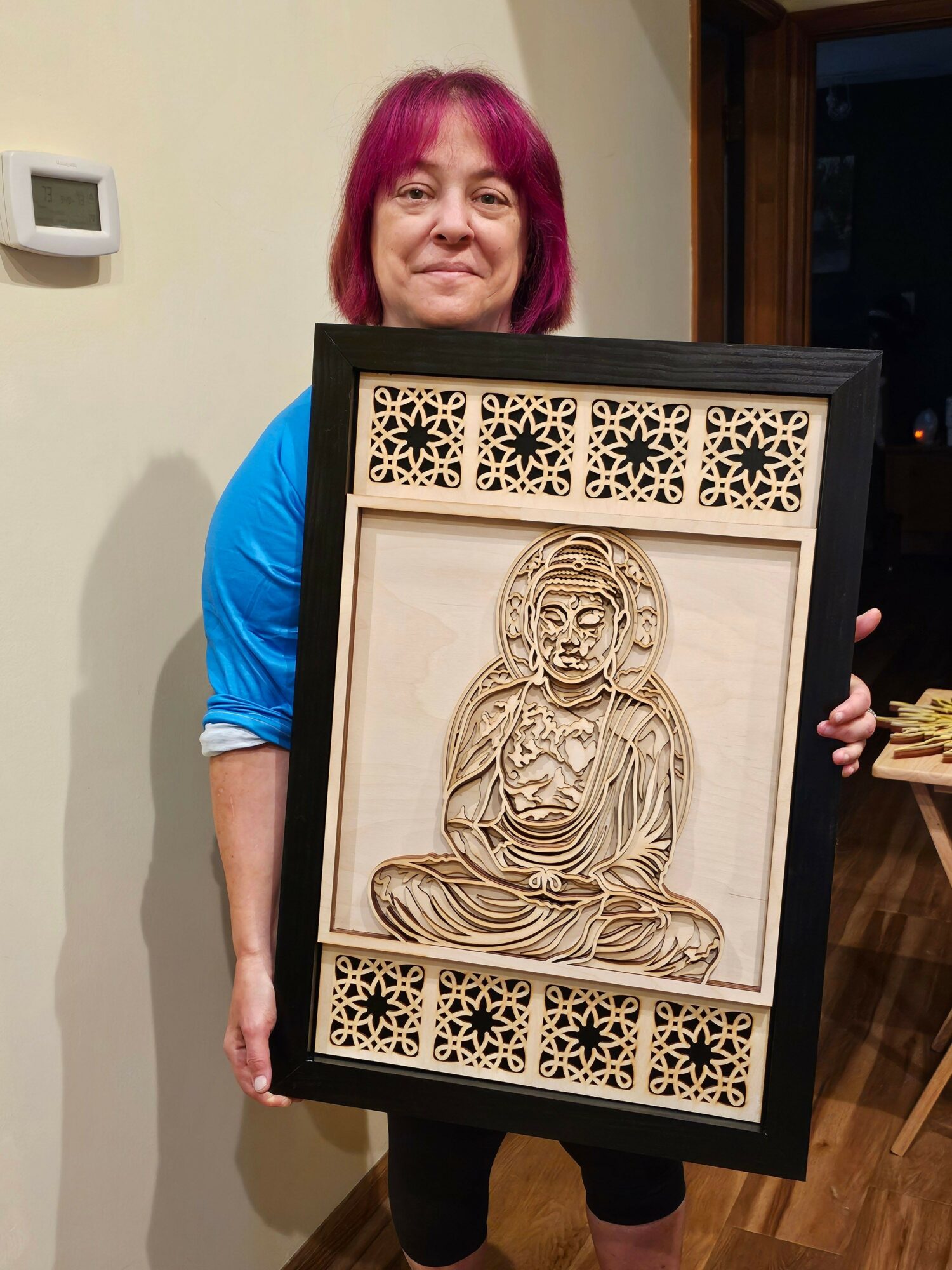 Woman with pink hair holding a framed artwork of a religious figure, with decorative patterns on sides, indoors.