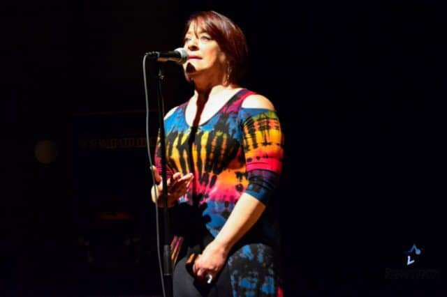 Woman with red hair singing into a microphone on stage, wearing a colorful patterned top, dark background