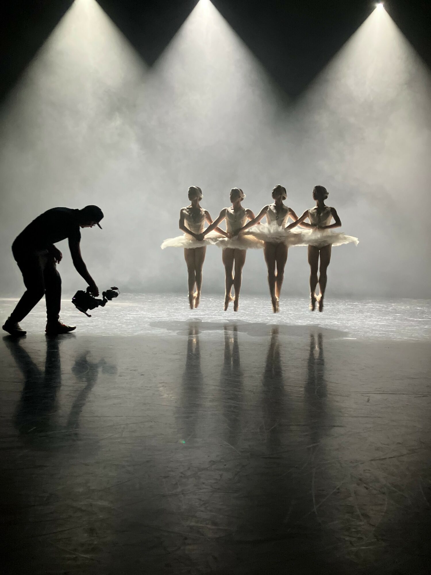 Four ballet dancers in tutus perform on stage with a photographer capturing the scene, backlit with bright lights and fog.