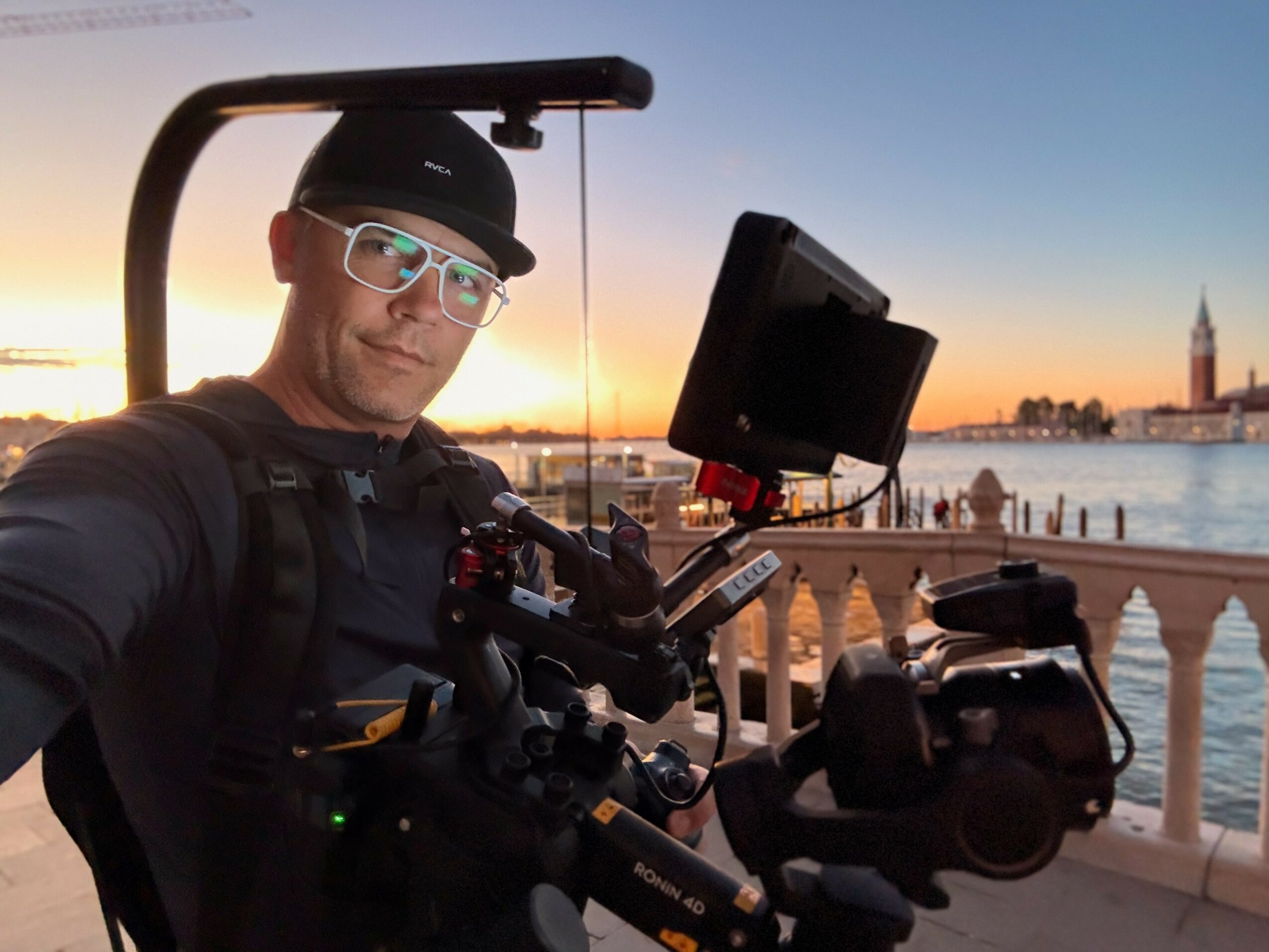 Person wearing glasses and a hat, operating camera equipment outdoors near water during sunset.