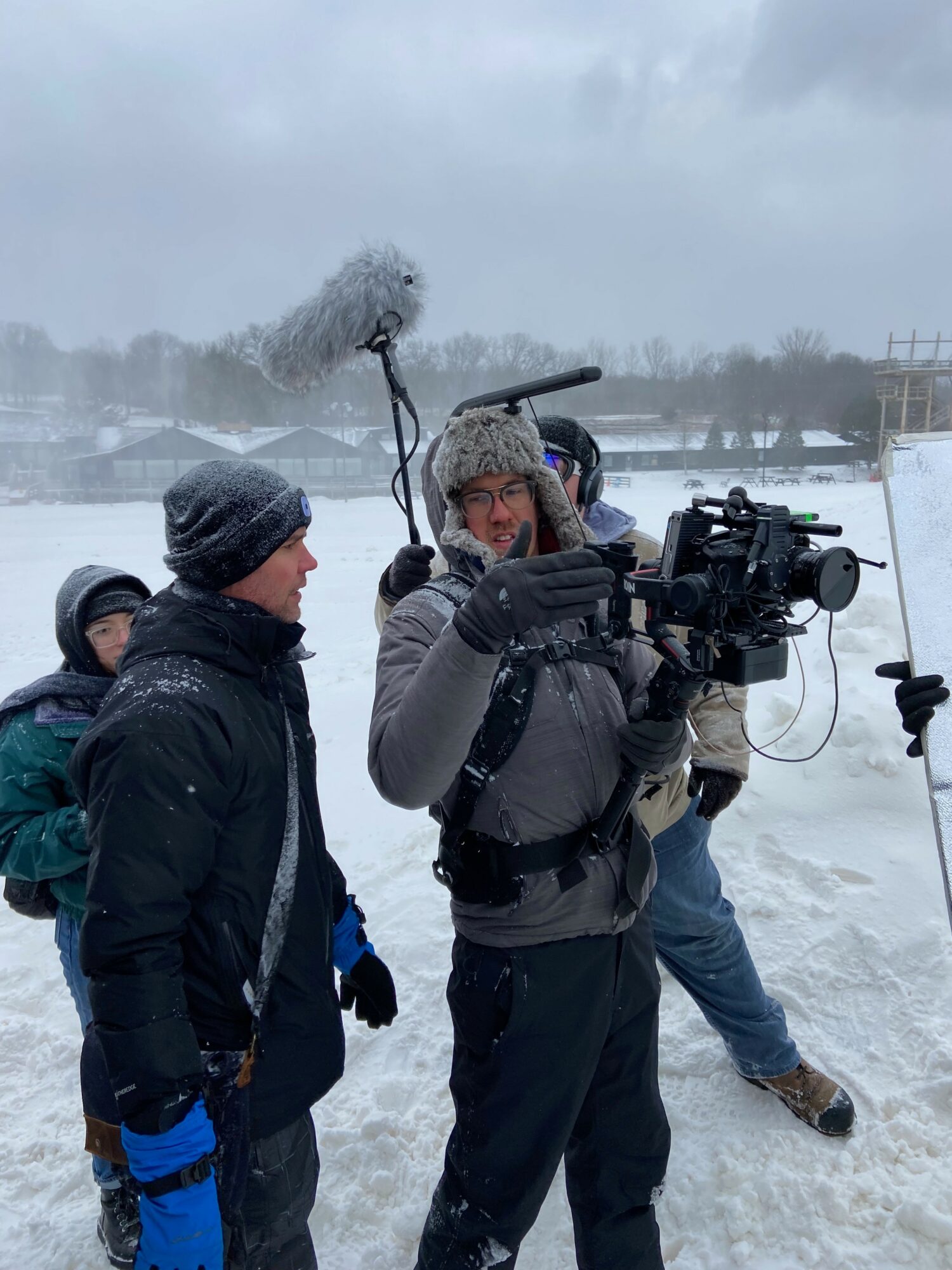 Group of people outdoors in snowy weather, one person holding a camera, others wearing winter clothing, snow-covered landscape