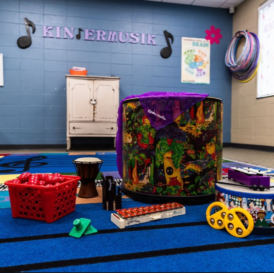 Children's music room with colorful instruments and decorations, blue wall, and a cabinet, labeled 'KINDERMUSIK'.
