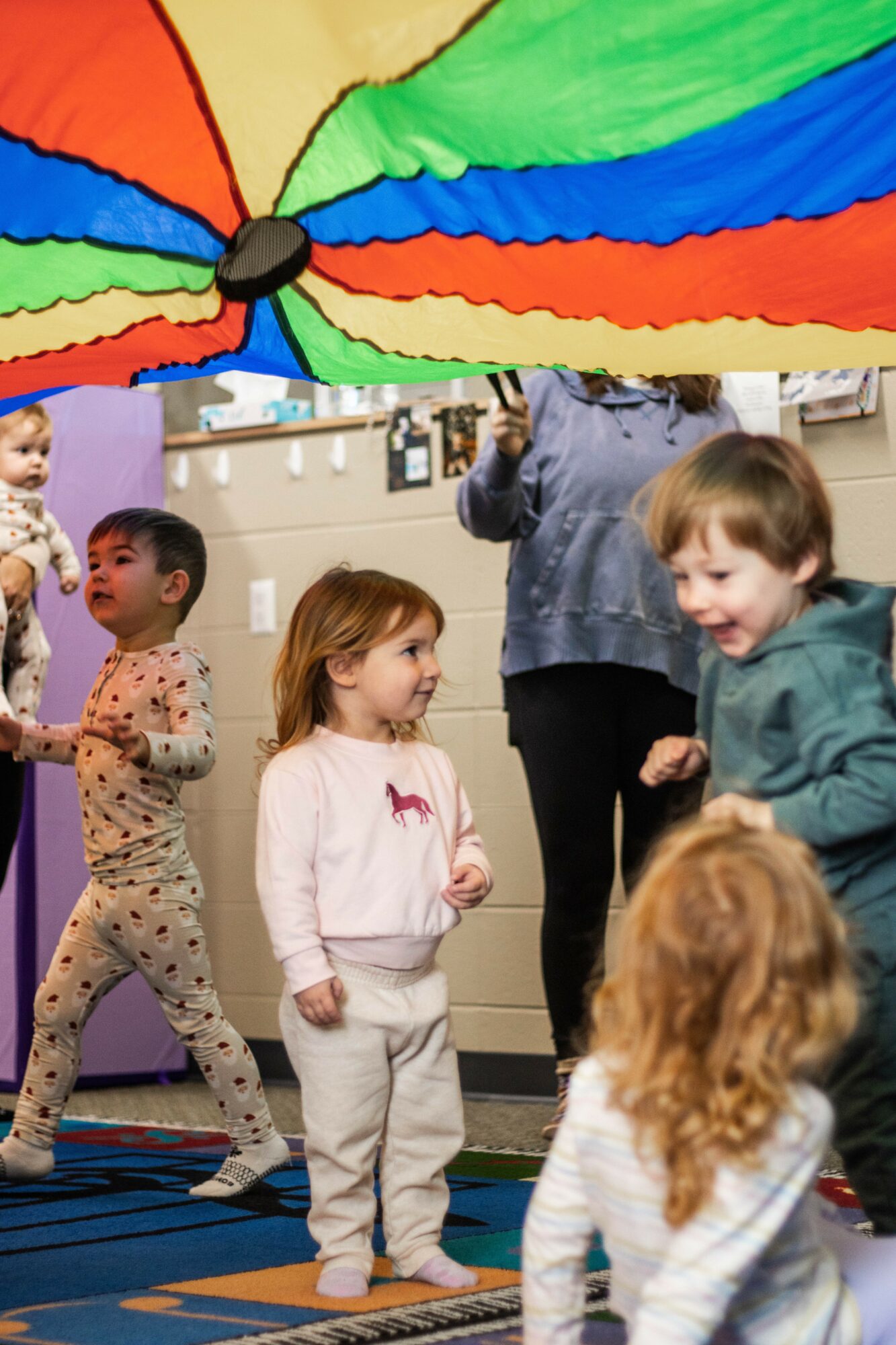 Children playing indoors under a colorful parachute held by an adult, with a patterned carpet and wall decorations.