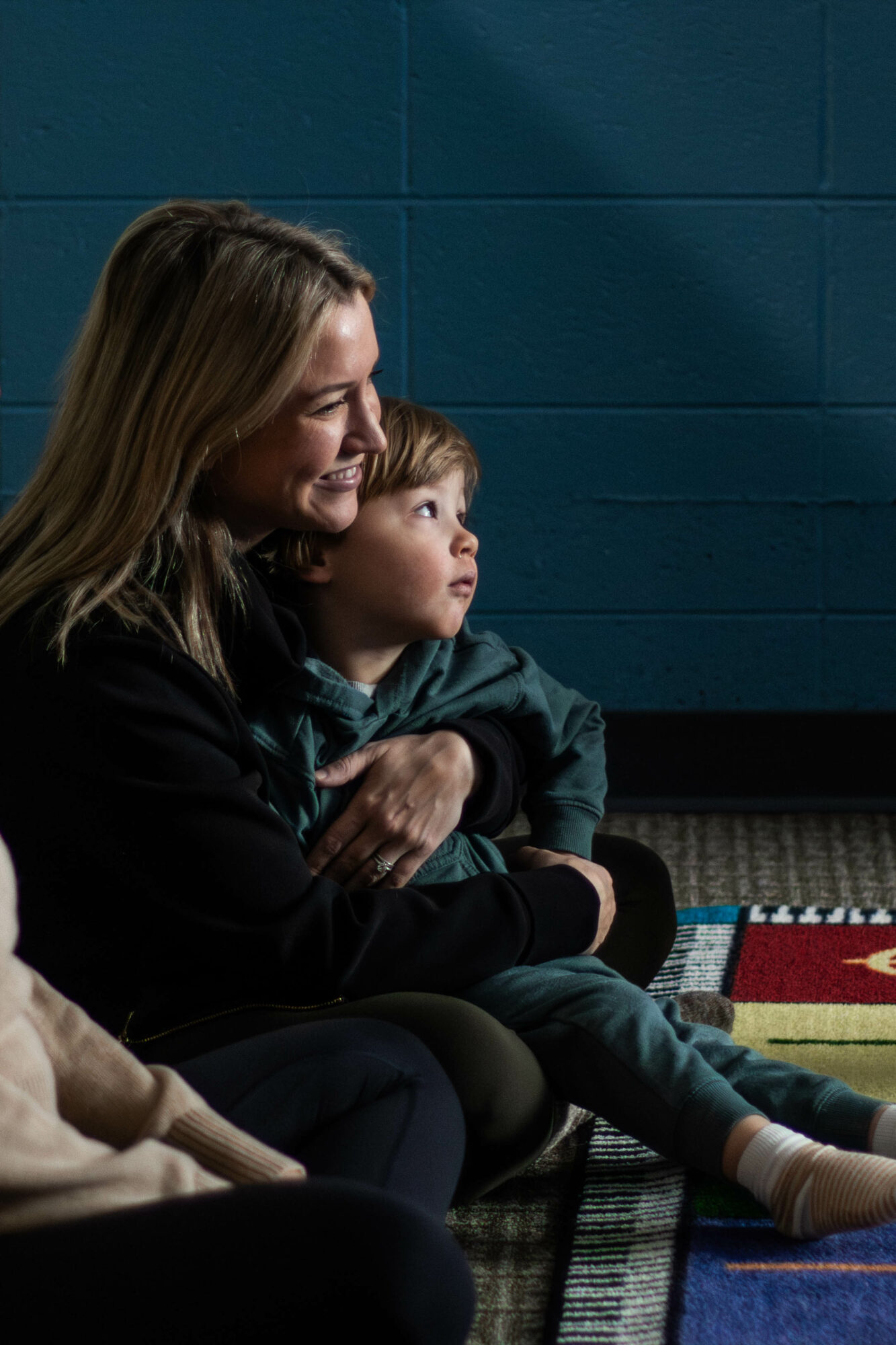 Woman and child sitting together, smiling, against a blue wall, with a colorful mat on the floor.