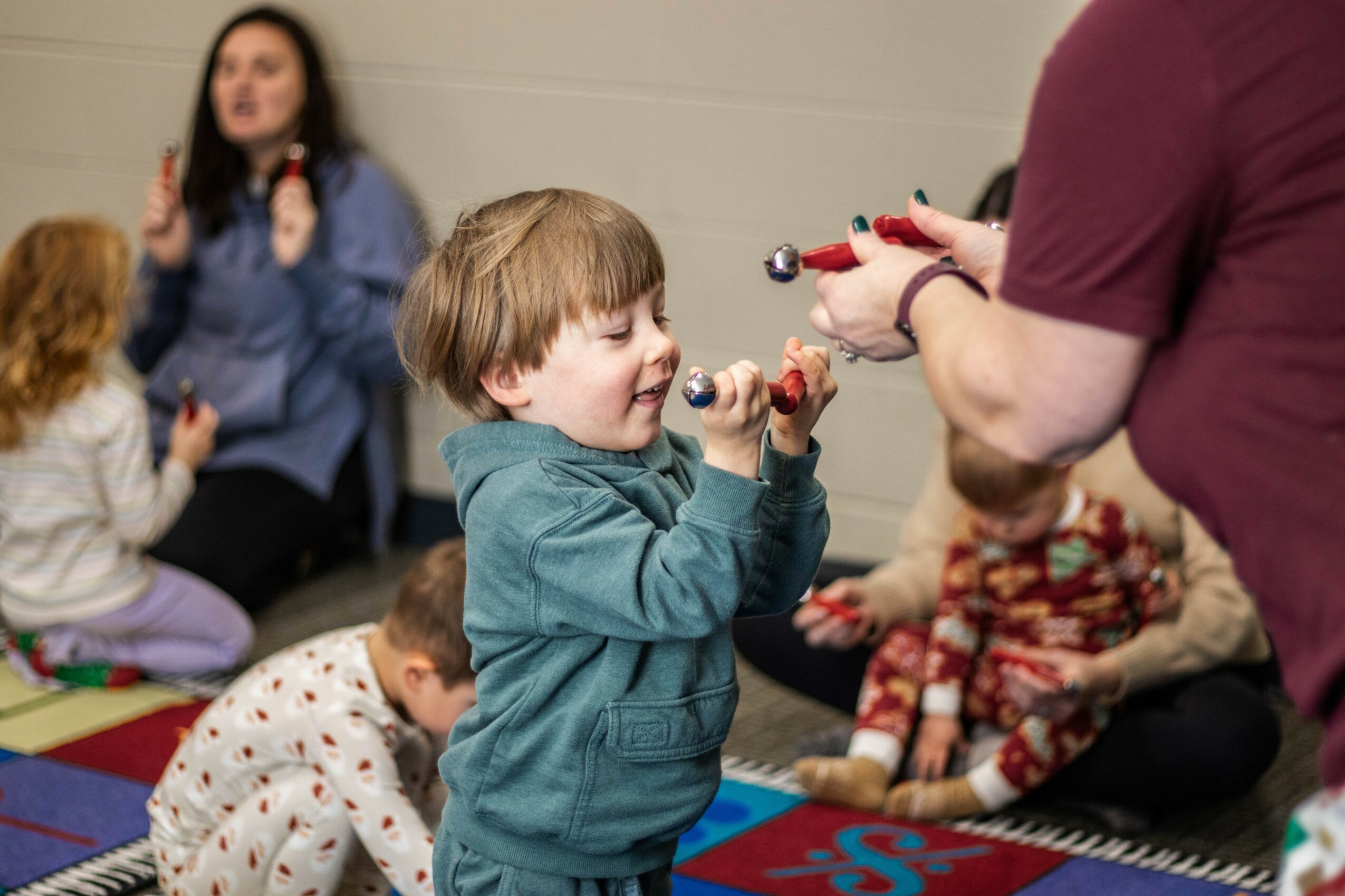 Child holding maracas, with adults and children sitting on mats in background, indoors, engaging in activity.