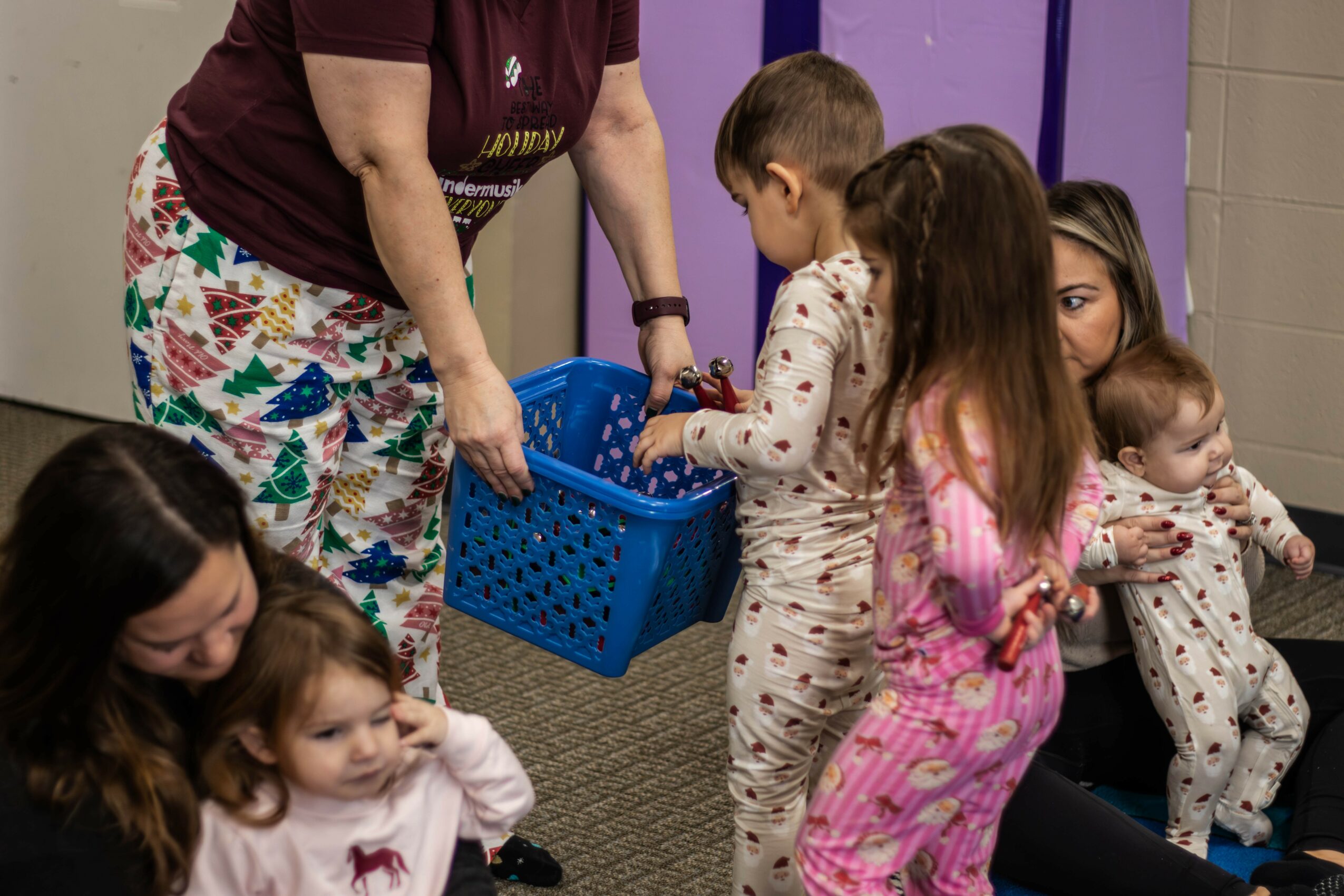 Group of children and adults gathered indoors, some children in pajamas, engaging in an activity with a person holding a blue basket.