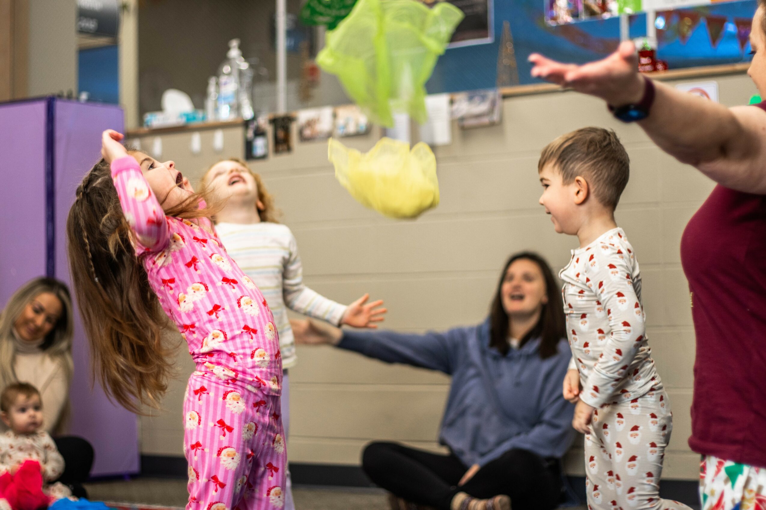 Children and adults playing with yellow tissue paper in a room with colorful decorations and shelves.