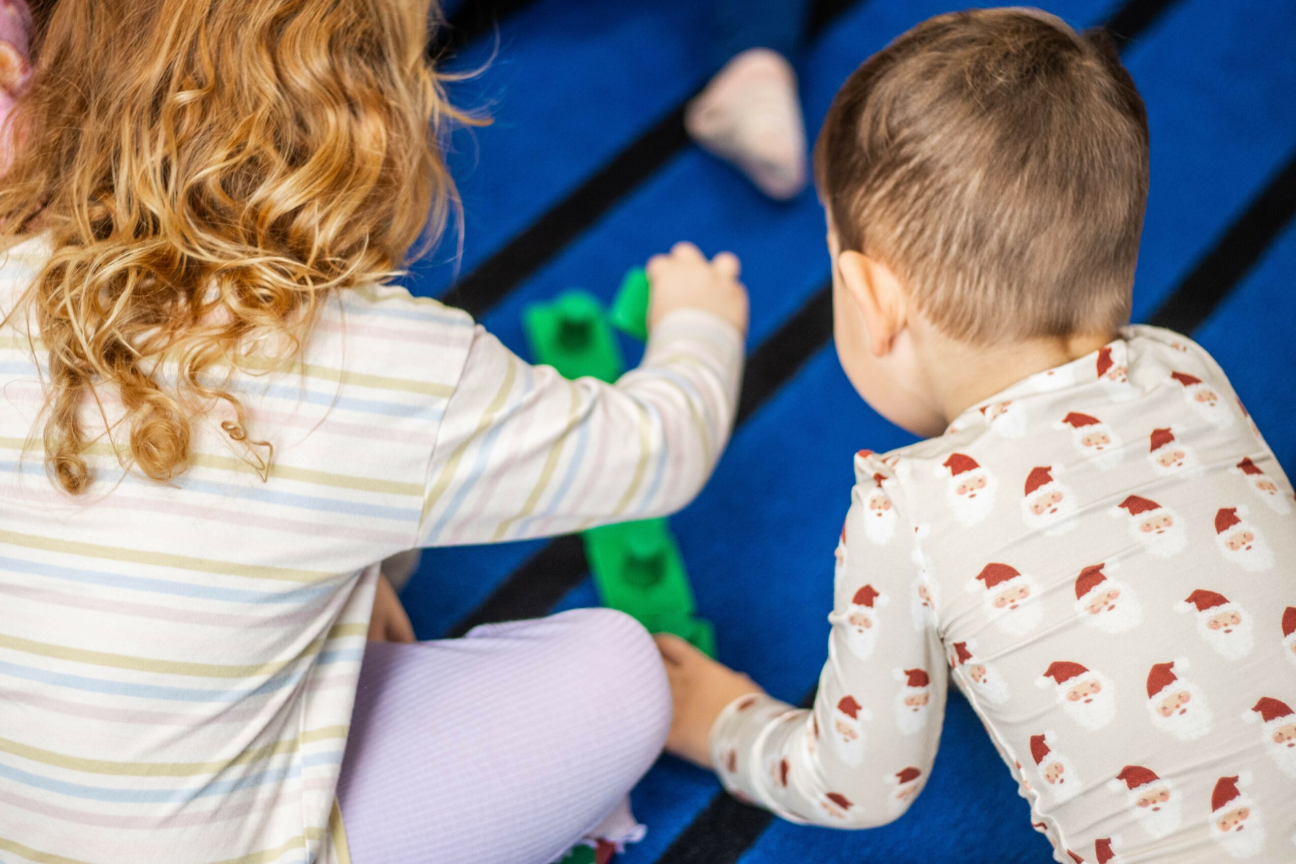 Two children playing with toys on a blue surface, seen from behind, one with curly hair and striped shirt, the other with short hair and patterned shirt.