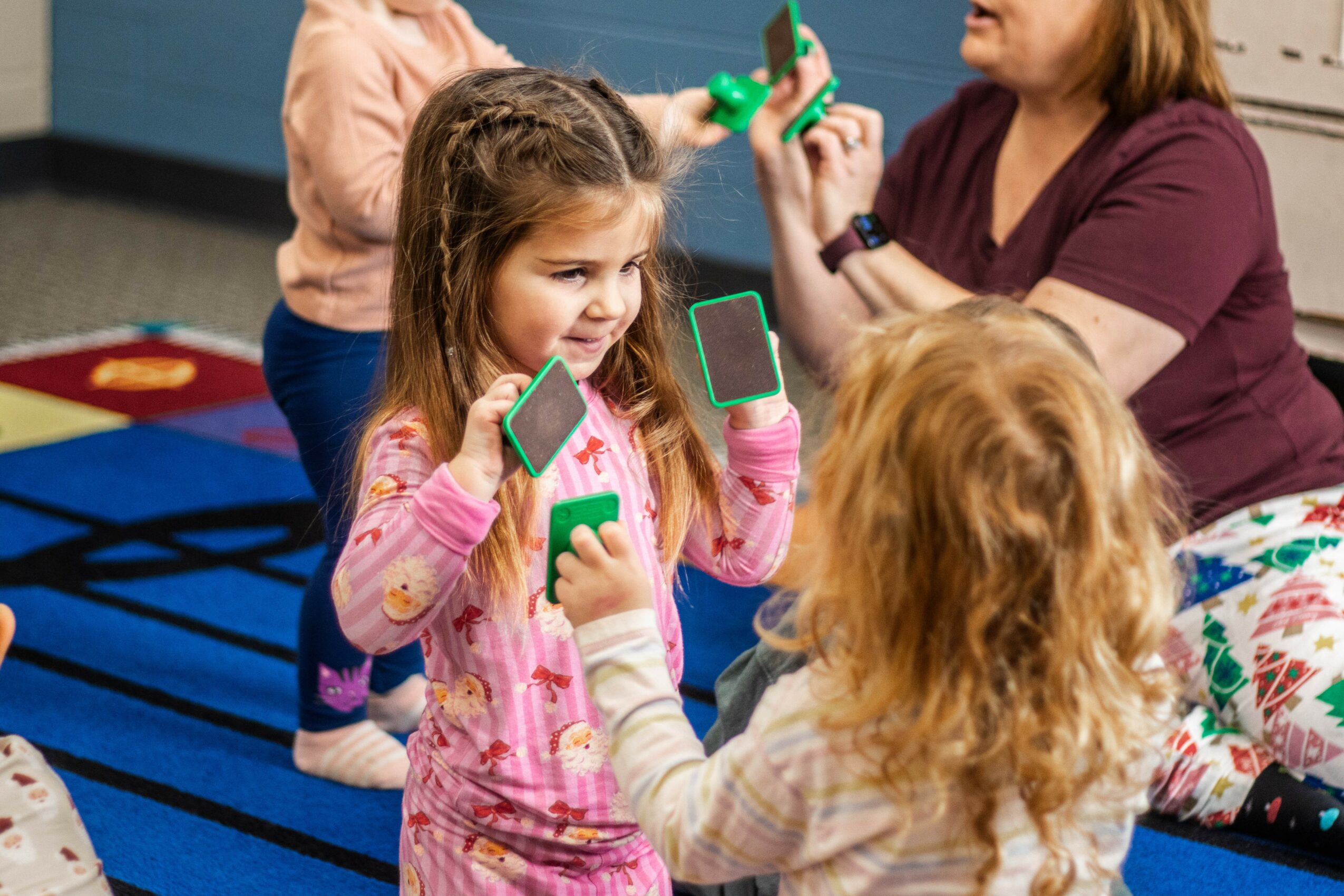 Two young girls with green toy phones talking, with an adult woman in the background, on a colorful play mat.