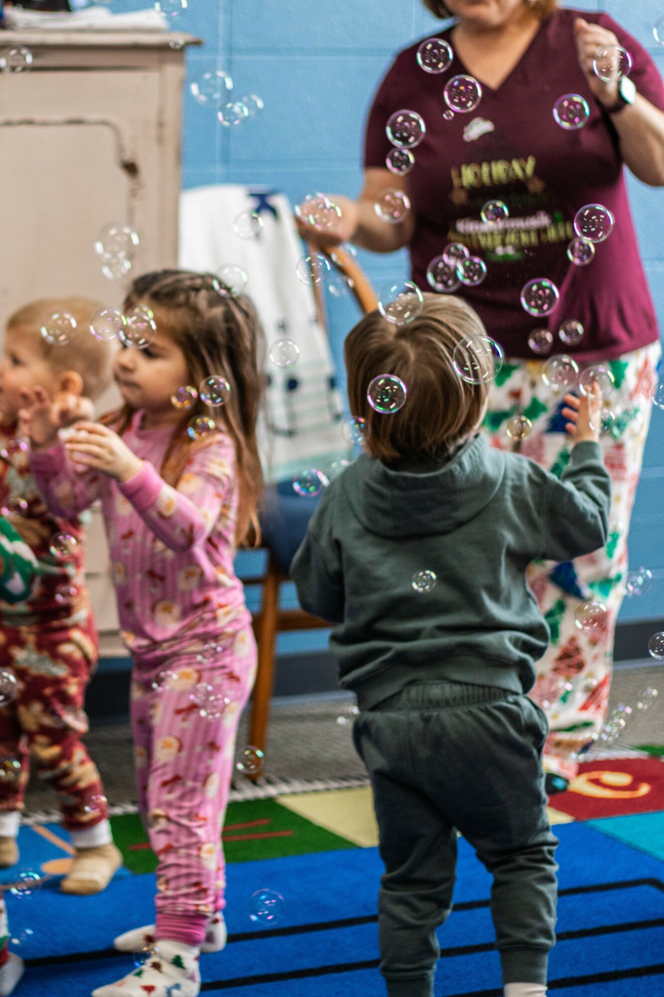 Children and an adult in a playroom with bubbles, colorful carpet, and furniture, engaging in play activities.