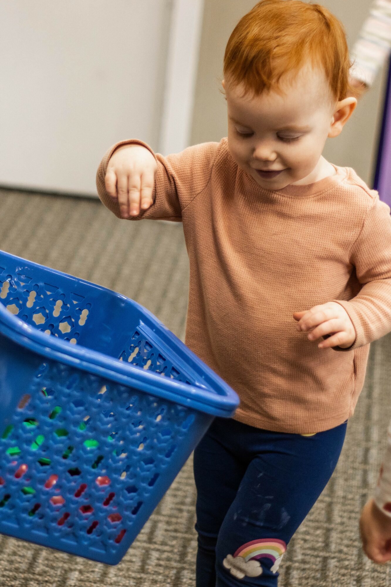 Child with red hair reaching into a blue laundry basket, wearing a peach shirt and dark pants.
