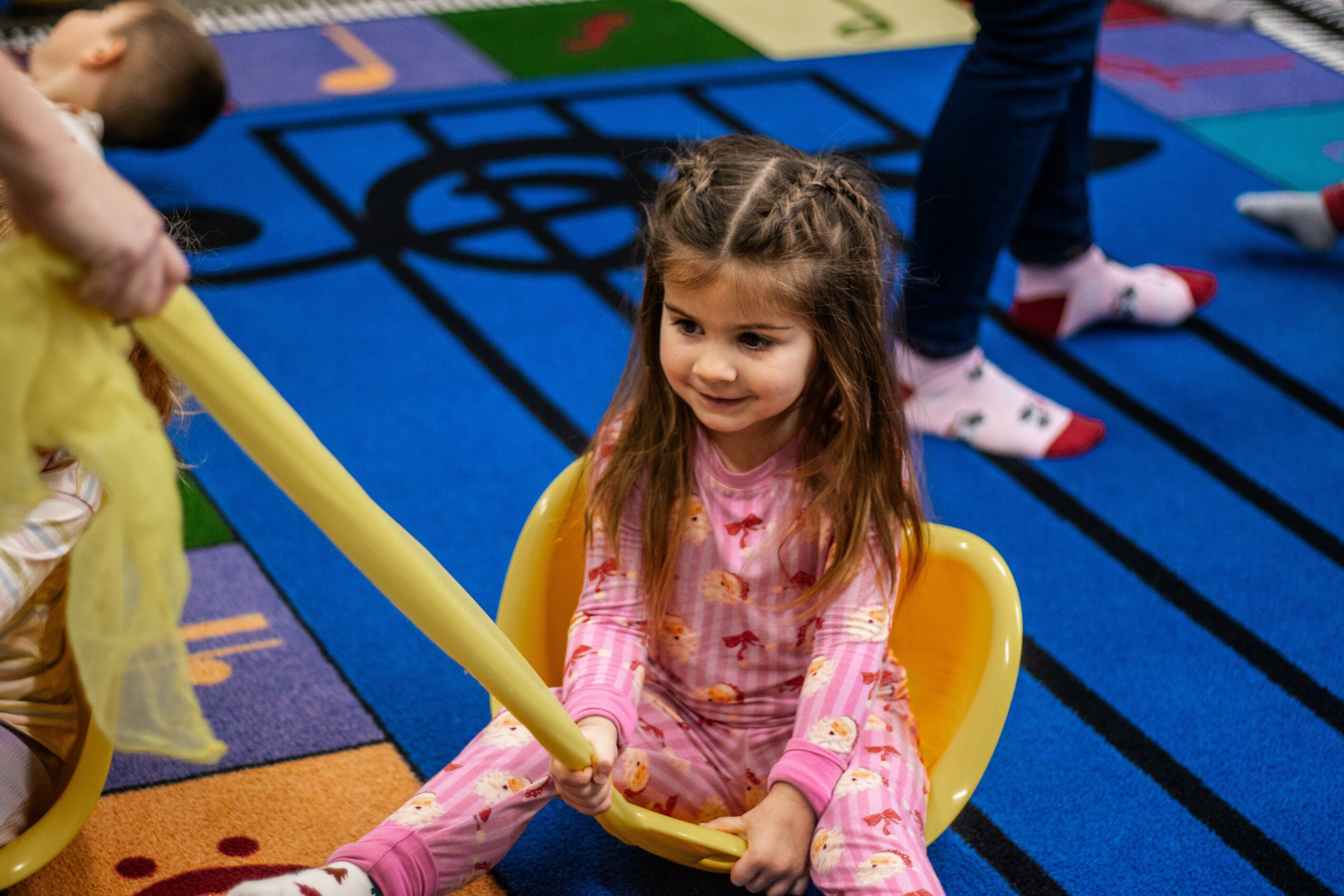 Young girl with long hair sitting in a yellow chair, holding a yellow balloon, on a colorful play mat.