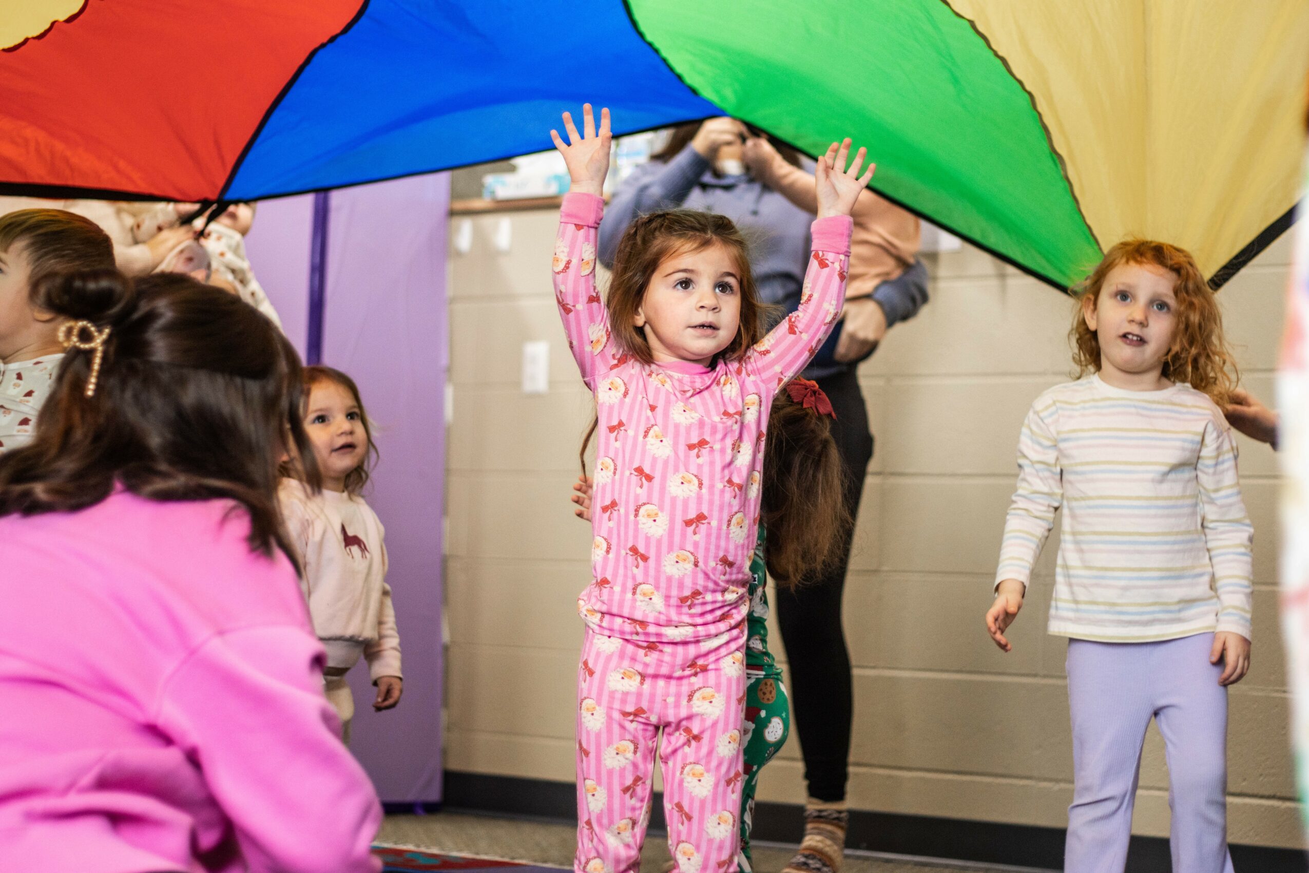 Children playing under a colorful parachute indoors, with one girl reaching up, surrounded by other kids and adults.