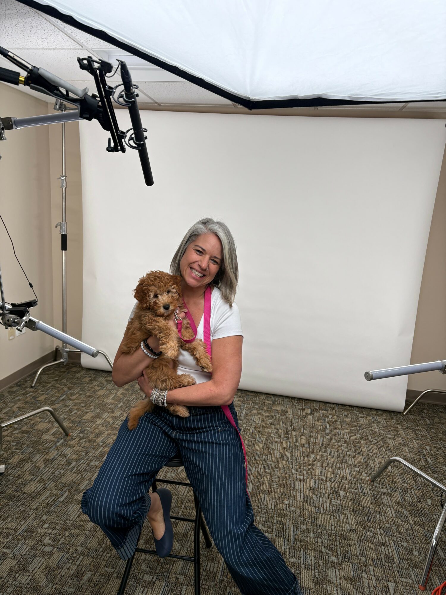 Woman holding a small curly-haired dog in a studio with a white backdrop and professional lighting equipment.